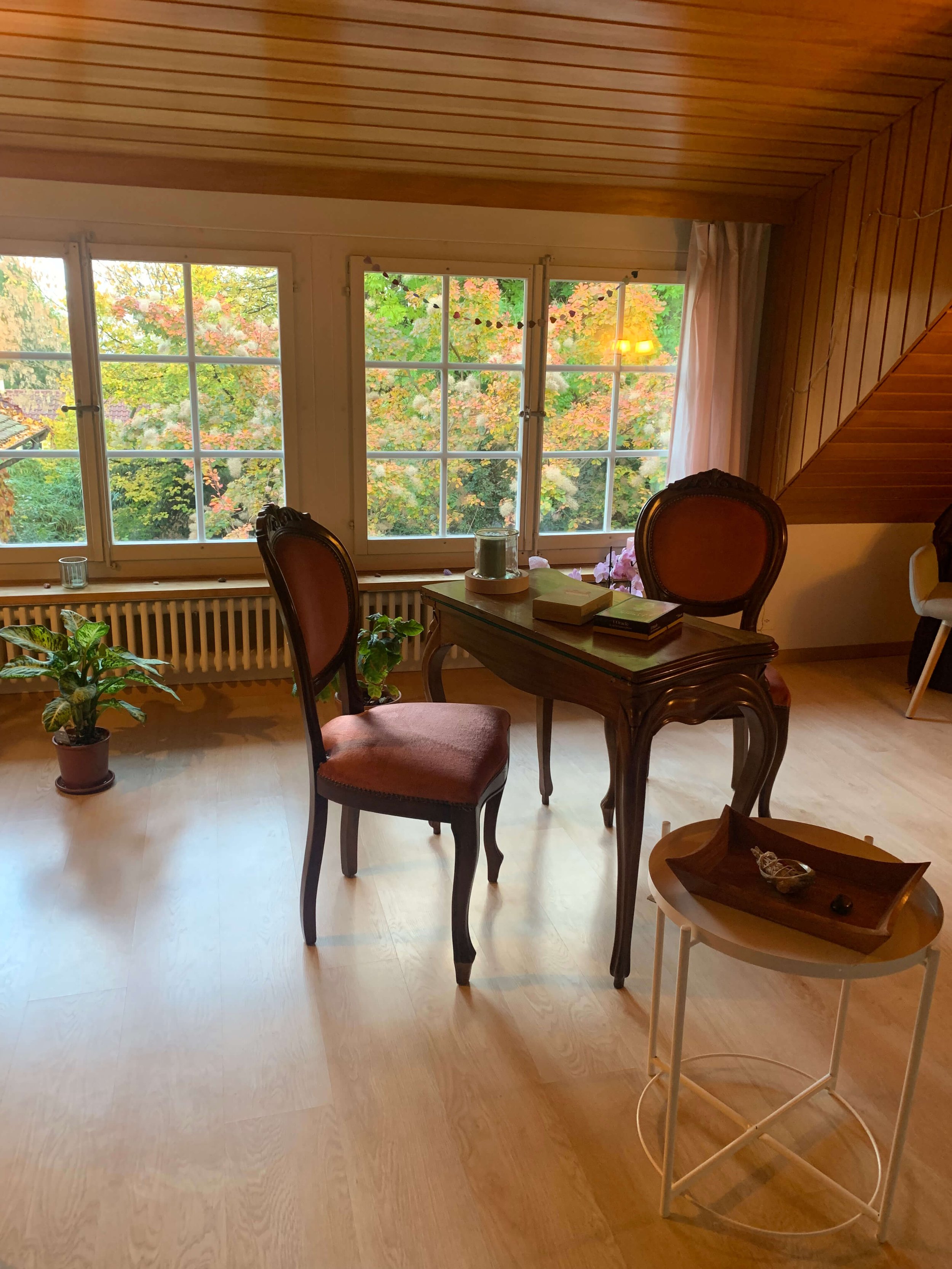 Intérieur d'une pièce avec trois fenêtres donnant sur un arbre aux feuilles colorées d'automne, un bureau en bois avec des livres et une bougie, deux chaises anciennes en velours et un petit meuble avec une assiette décorative.