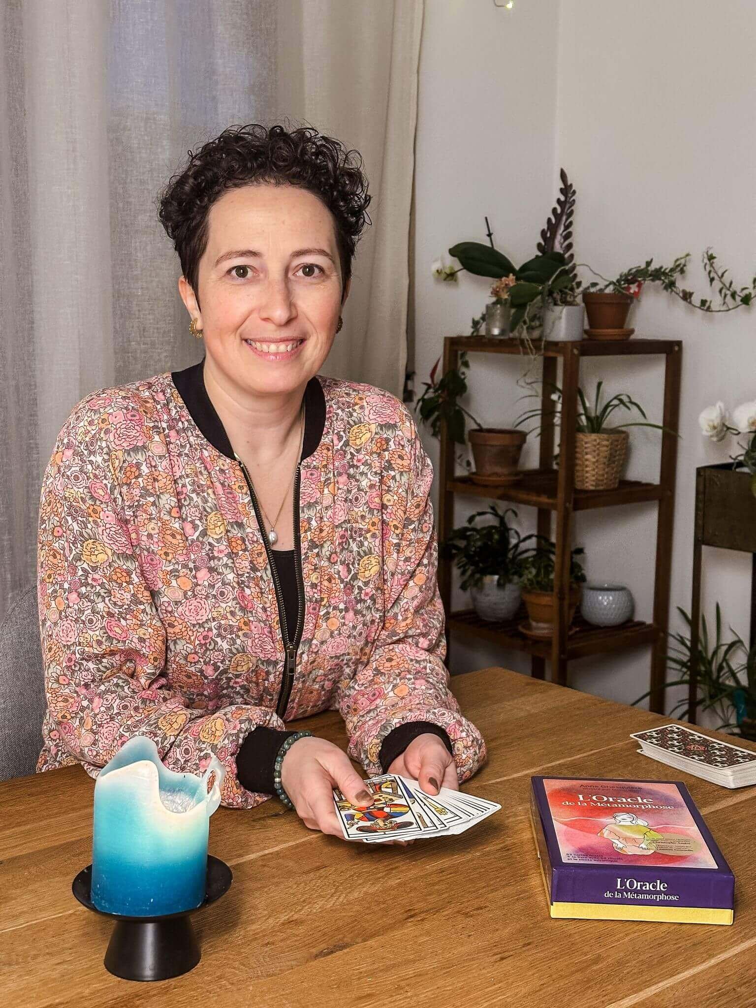 Une femme souriante tient un jeu de cartes devant un livre intitulé 'L'Oracle de la Métamorphose' sur un bureau en bois, avec une bougie bleue à côté.