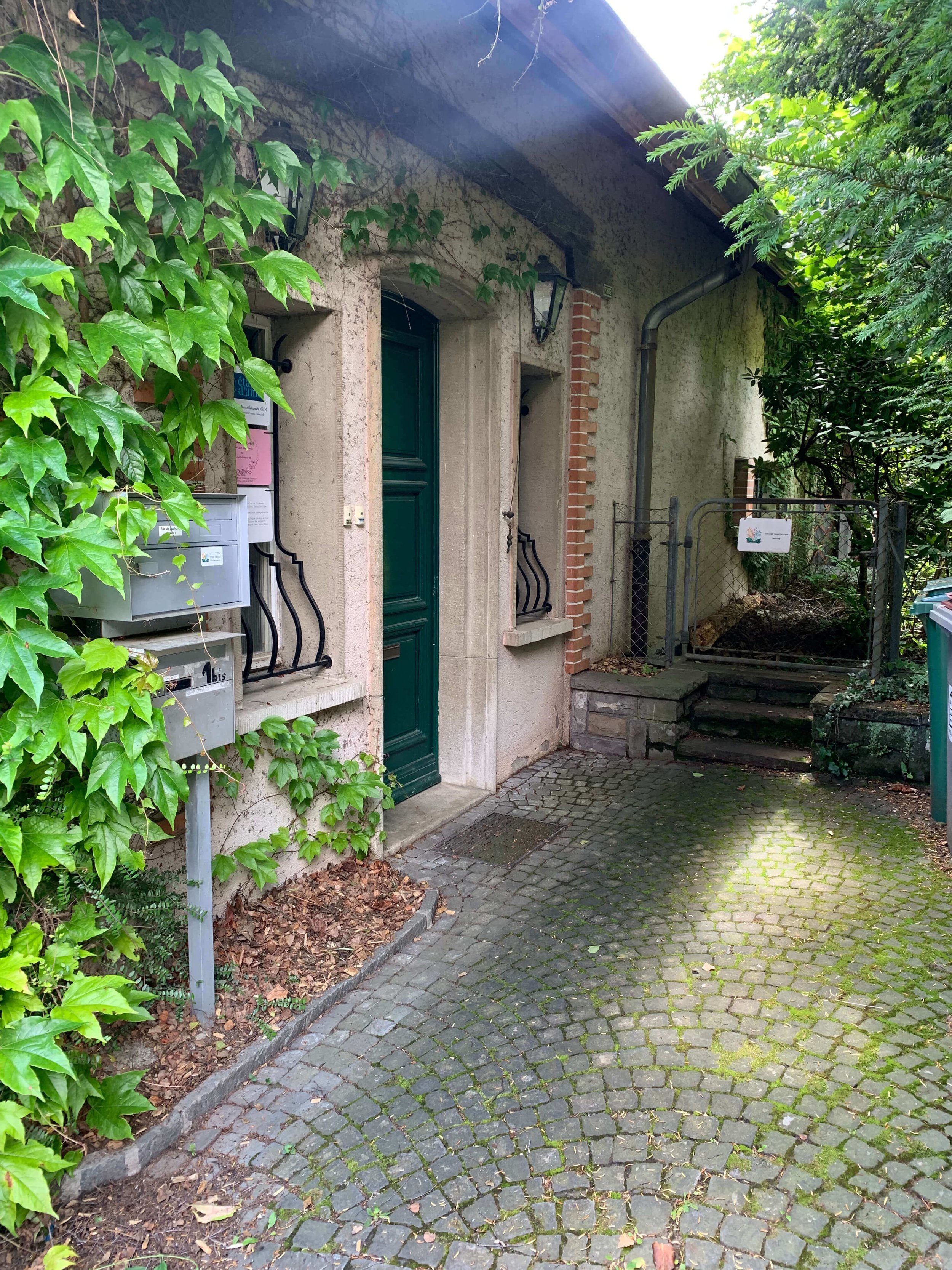 Allée pavée menant à une porte verte d'une maison en pierre, entourée de végétation et de feuillage vert, avec escaliers en pierre et clôture en métal.