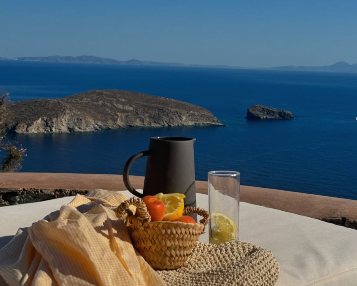 A table with a basket of fruit, a black pitcher, a glass of water with lemon, and some cloths, set against a scenic view of the ocean and islands in the background.