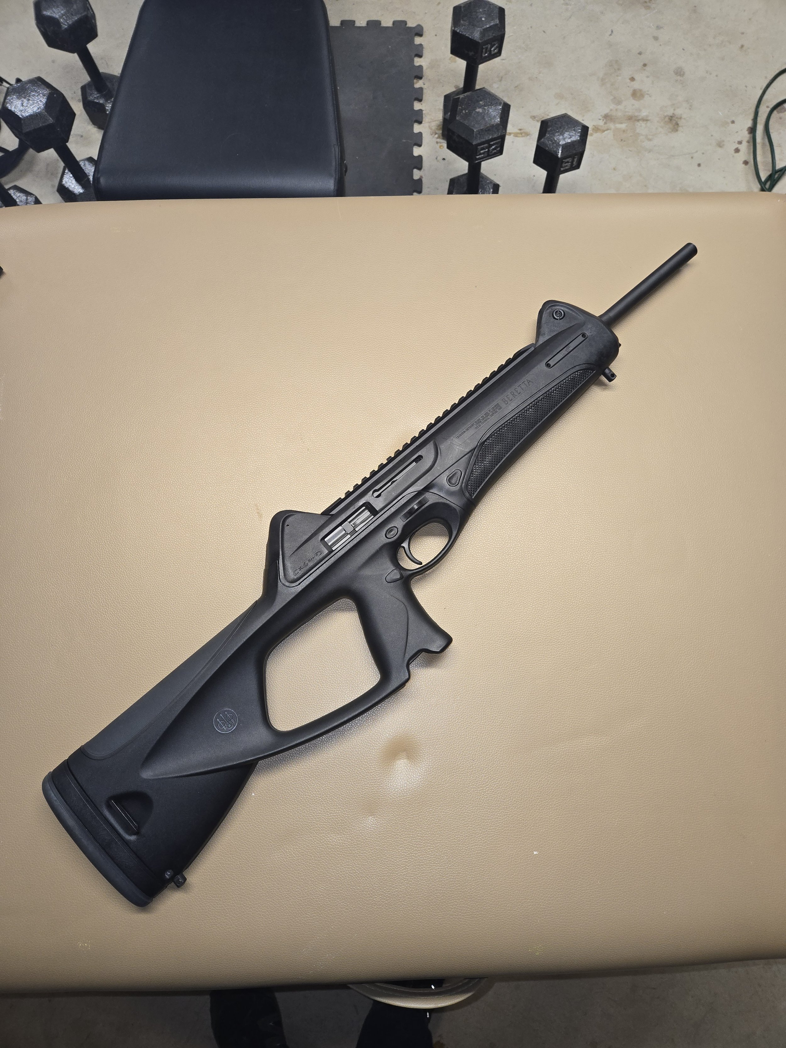 A black semi-automatic rifle with a synthetic stock rests on a beige table. In the background, there are black dumbbells and a black workout bench.