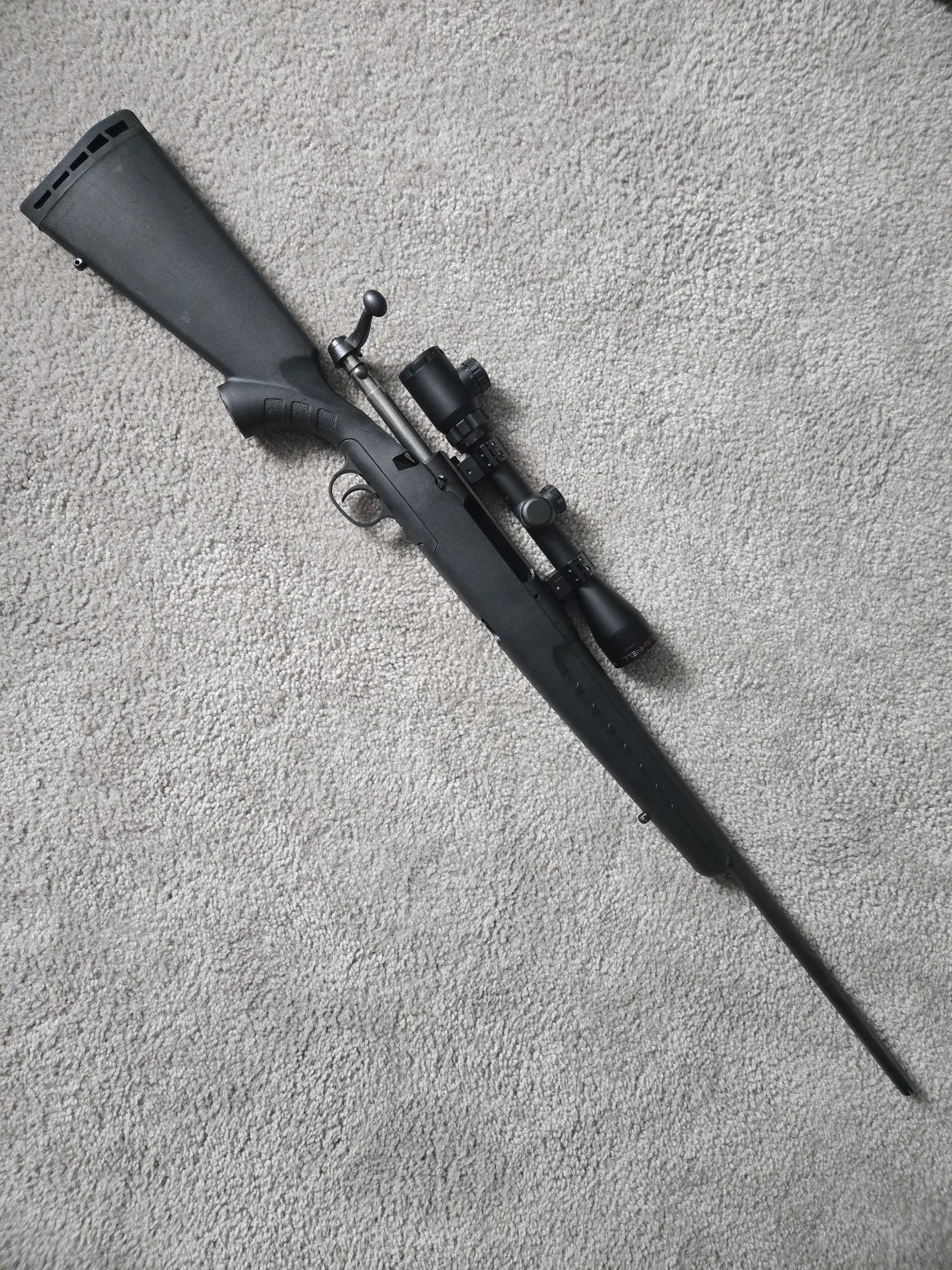 A black rifle with a scope, lying on a beige carpet.