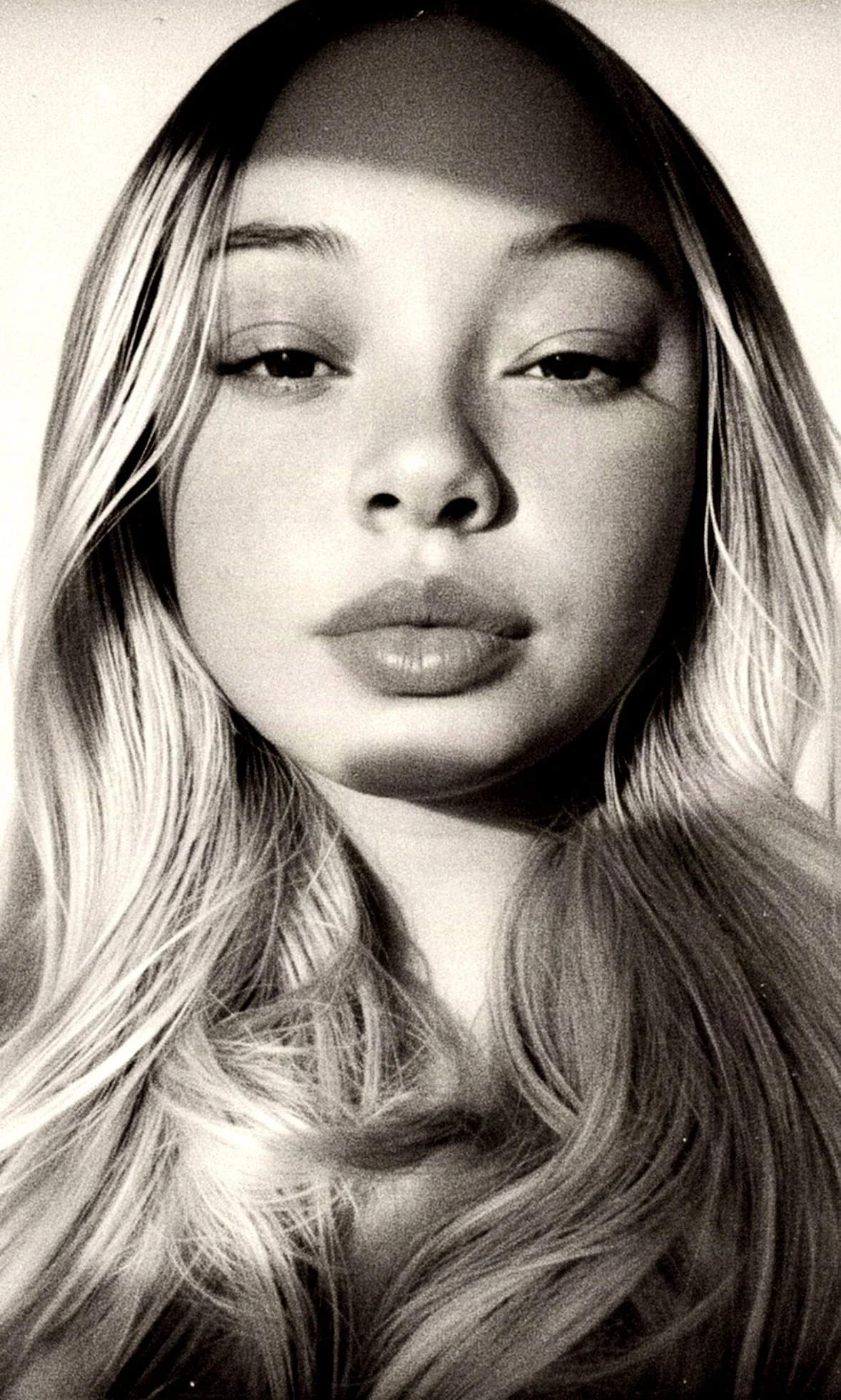 A black and white close-up photograph of a woman's face with long, wavy hair, partly hair-covered face, with a neutral expression.