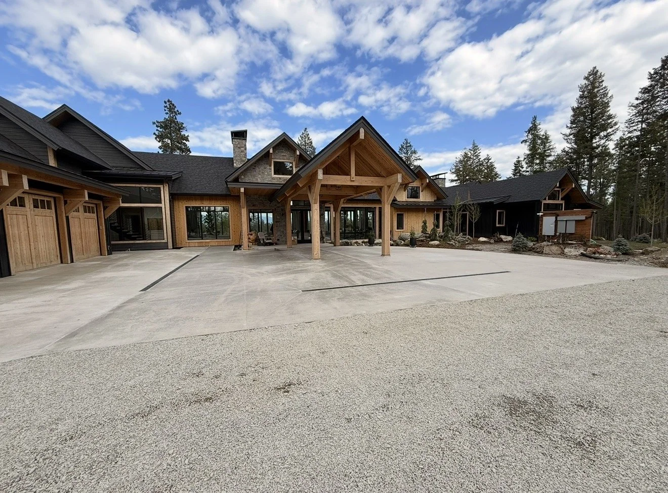 Large, newly constructed house with a mix of wood and stone exterior, multiple garage doors, and a prominent covered entrance supported by wooden beams. The house is set in a wooded area with a circular driveway and a gravel surrounding driveway, under a partly cloudy sky.