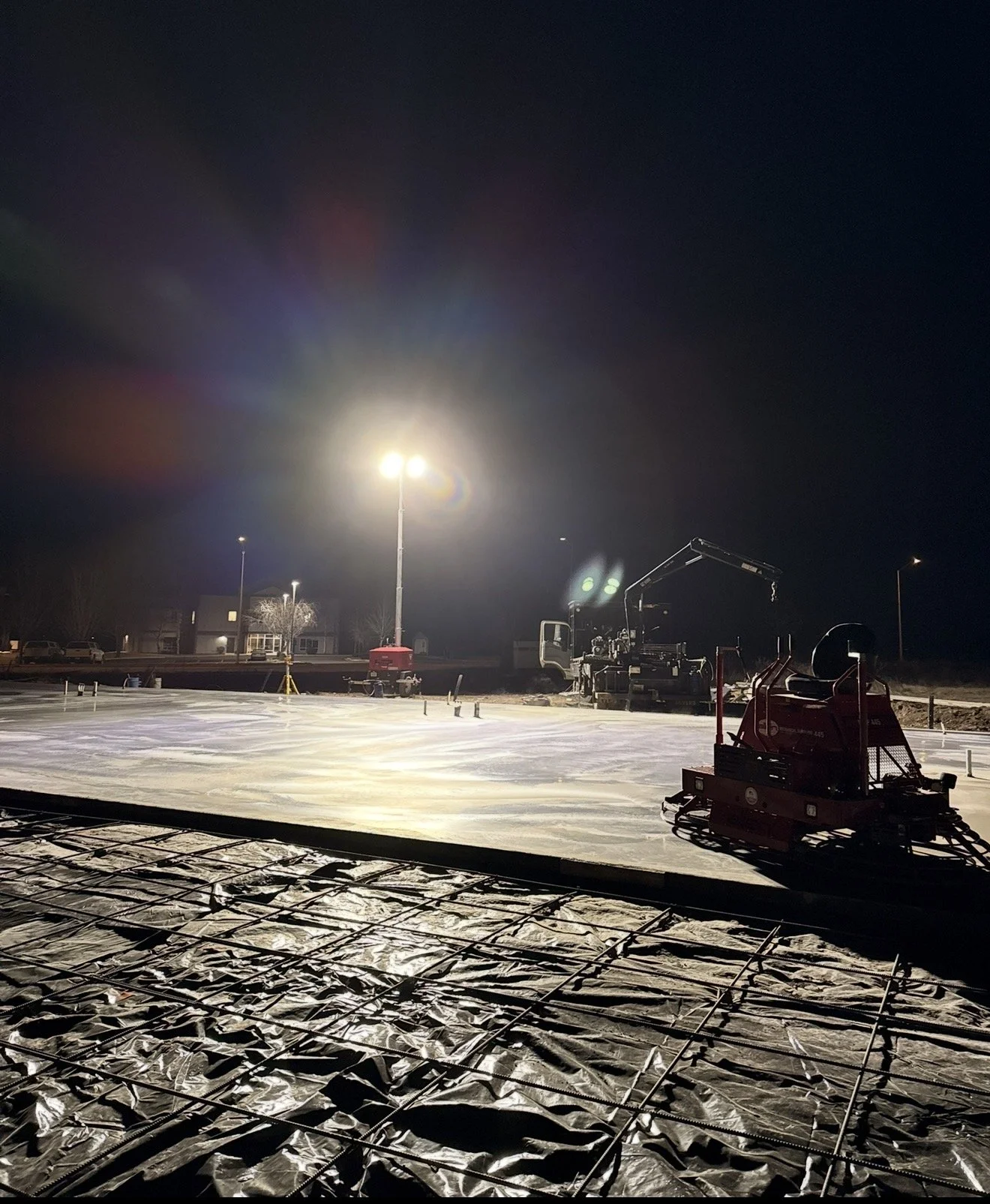 Nighttime construction site illuminated by bright floodlights, with machinery and rebar on the ground.