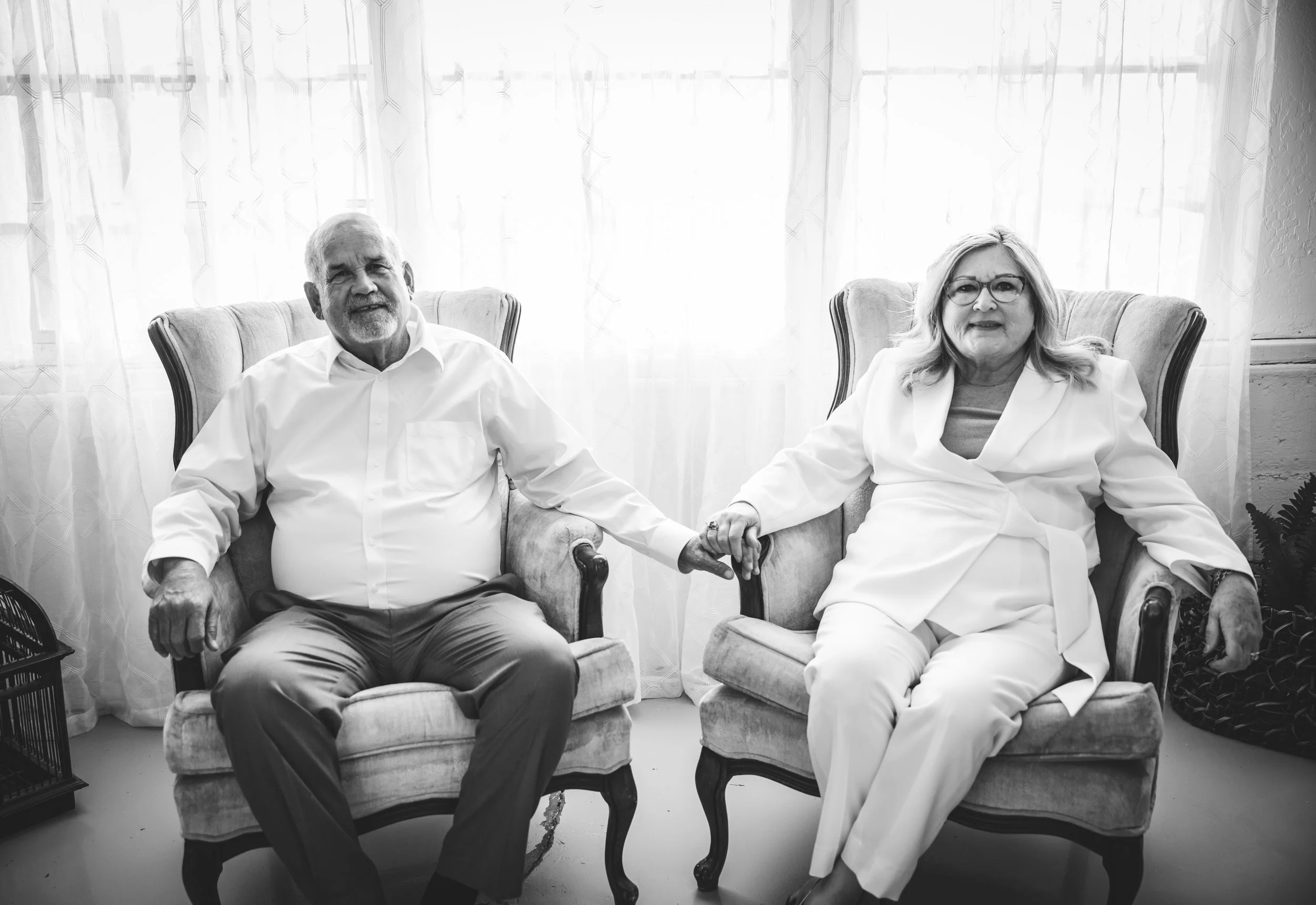 An elderly man and woman sitting on vintage armchairs holding hands in front of a sheer curtain. The man has a beard, is wearing a white shirt and dark pants. The woman has glasses, is wearing a light-colored blazer and pants, and has shoulder-length