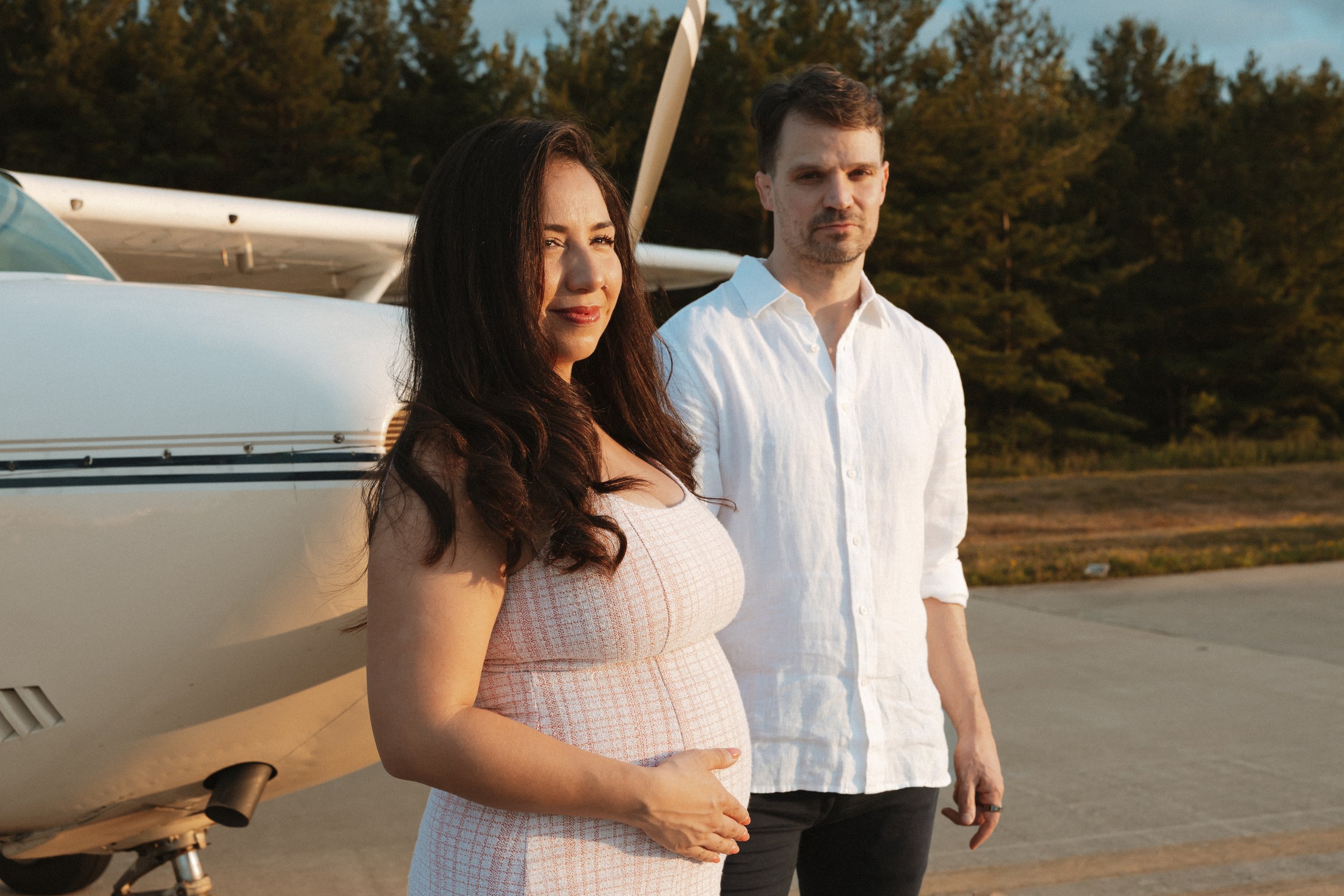 A pregnant woman with long dark hair in a pink and white dress standing next to a man with short brown hair in a white shirt near a small airplane on a runway during sunset.