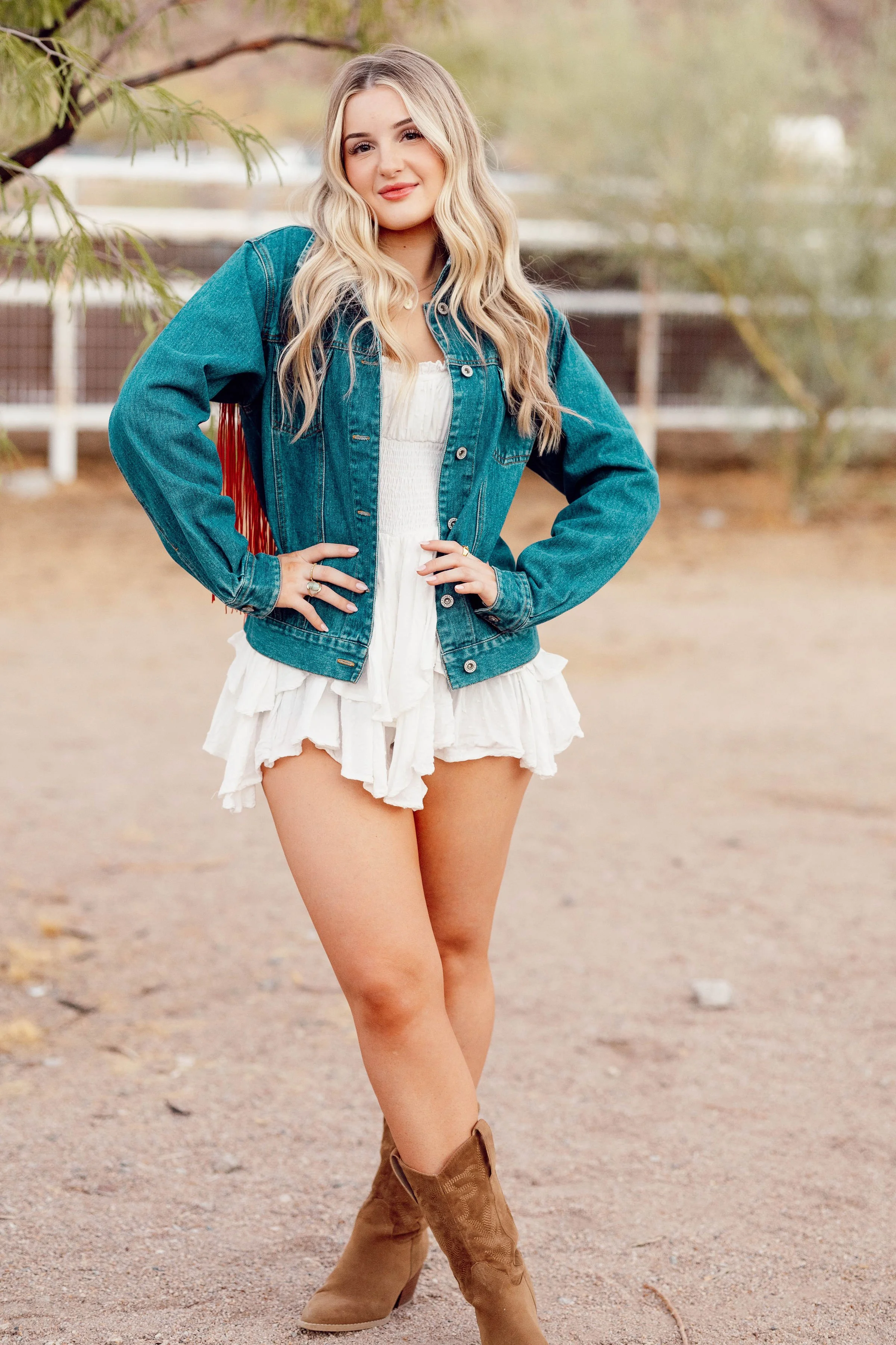 A young woman with long blond hair wearing a denim jacket, a white ruffled romper, and cowboy boots standing outdoors in a natural setting with a fence and trees in the background.