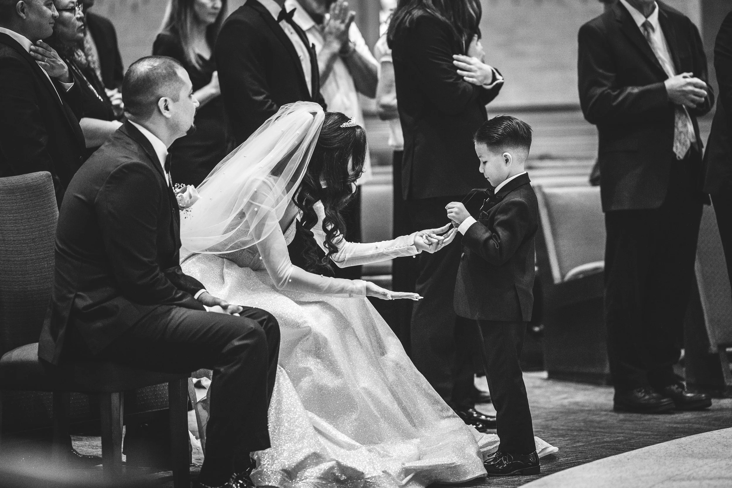 A bride in a wedding dress and veil interacts with a young boy in a tuxedo during a wedding ceremony, surrounded by seated and standing guests in formal attire.