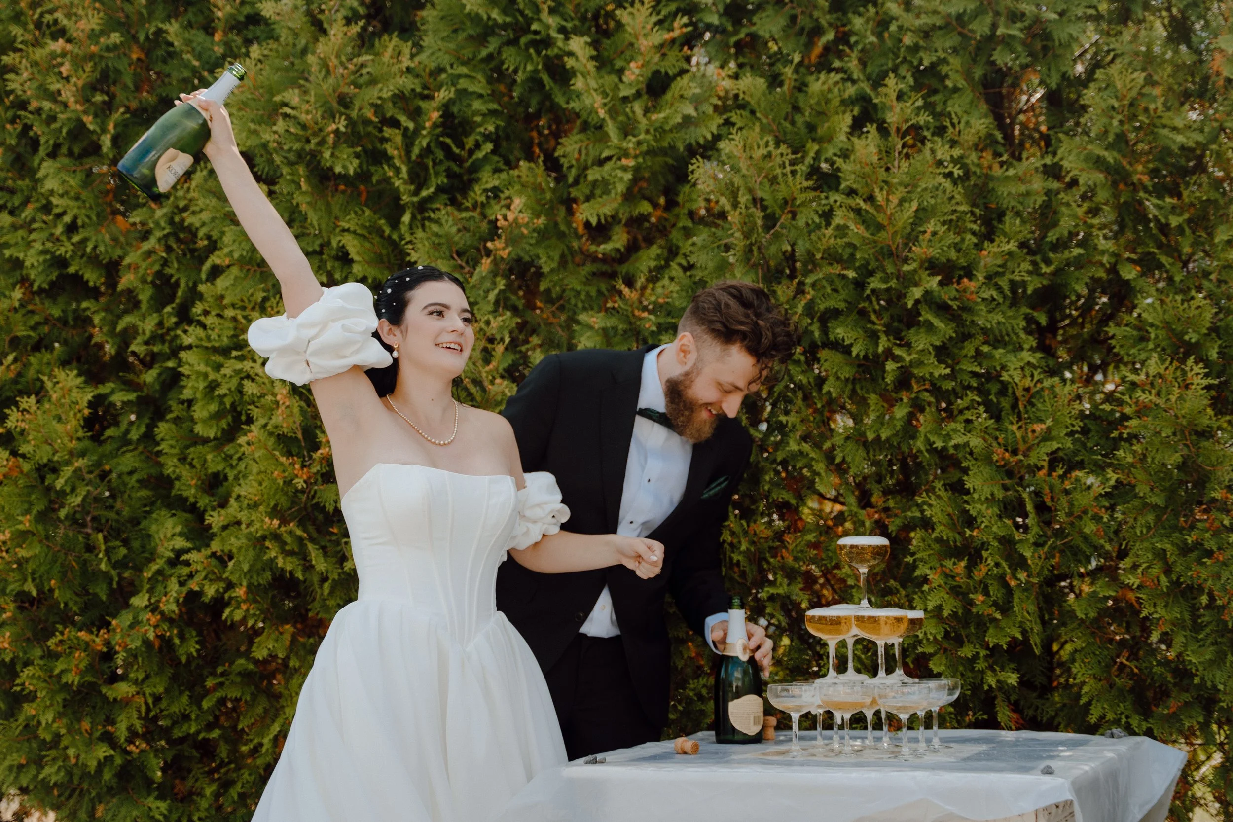 Bride and groom celebrating a wedding outdoors, with the bride holding a champagne bottle in the air and the groom smiling, as they stand next to a champagne tower on a table. Wedding Video by Lasting Legacy Films