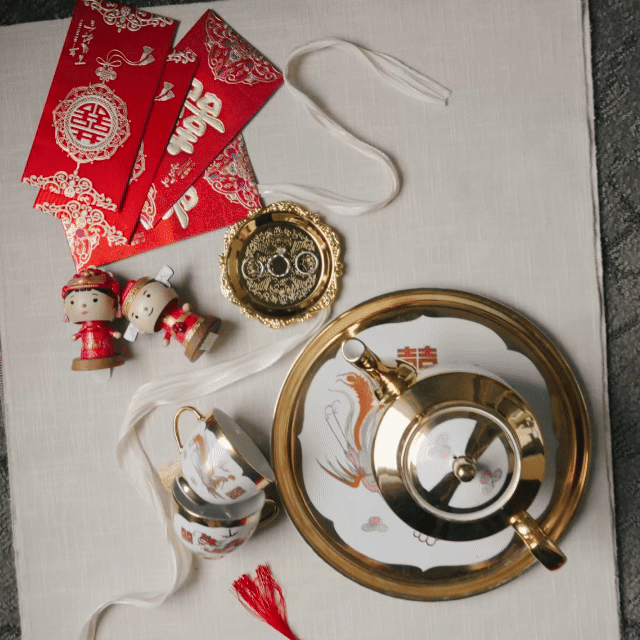 Wedding Video Chinese Wedding. Decorative items for a Chinese celebration on a white surface, including red envelopes,  a gold-colored teapot, two small cups, and a gold and white tray with a Chinese character and a dragon design. Wedding Venue 