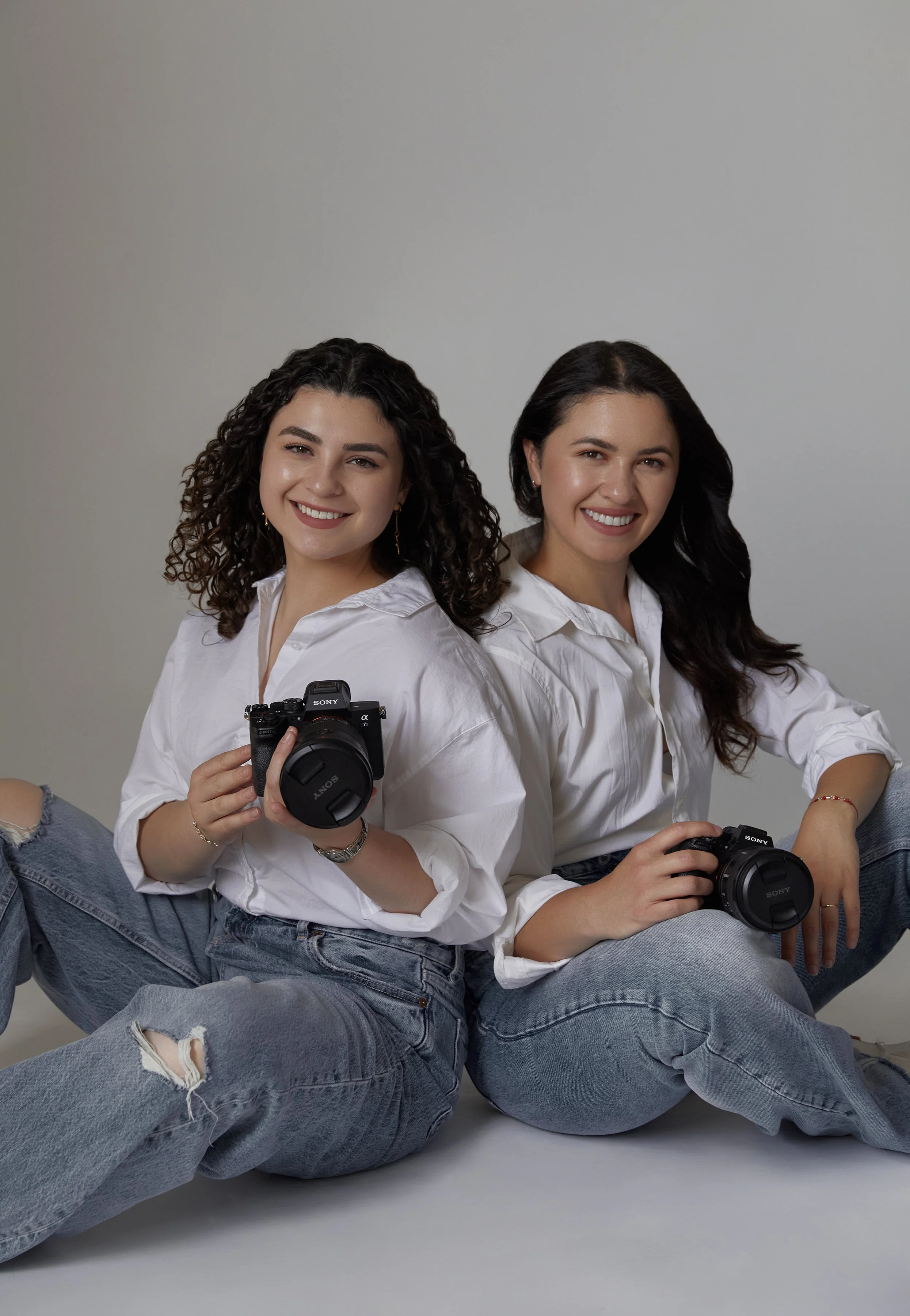 Two women sitting on the floor, smiling, each holding a camera; one camera pointed towards the viewer, the other towards the side, both wearing white shirts and ripped jeans, against a plain gray background. Wedding Videographers smiling