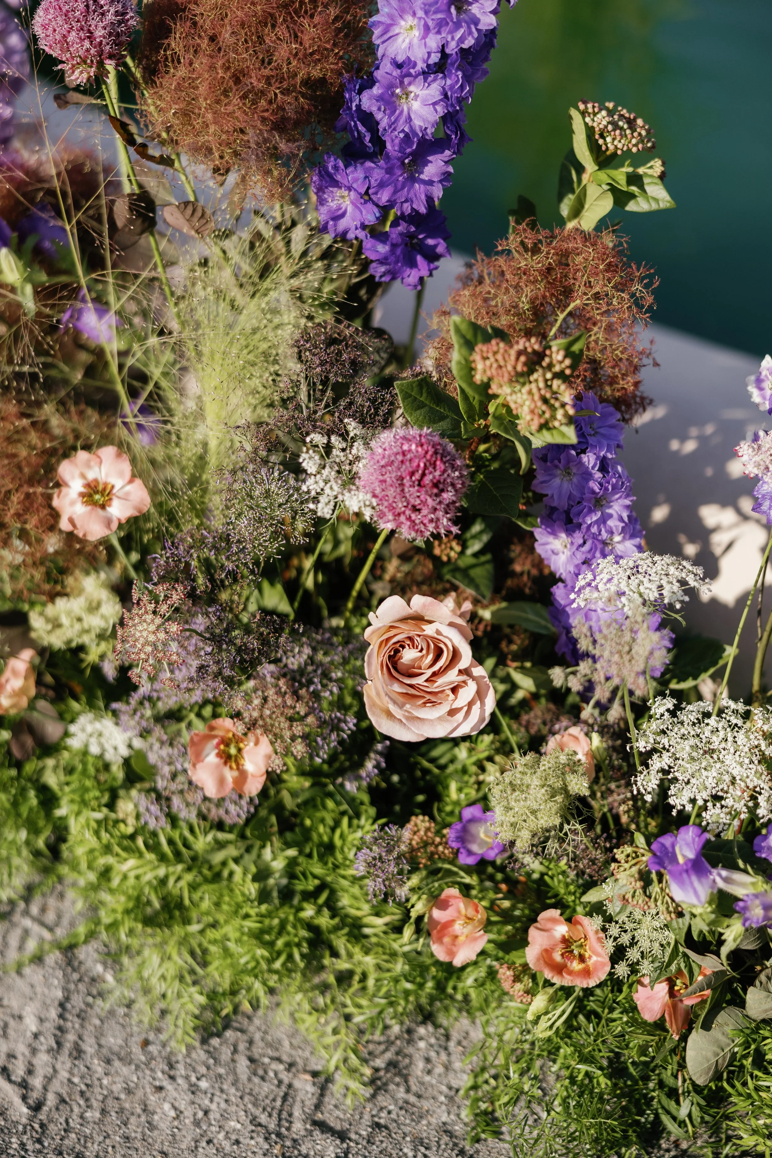 A colorful arrangement of various flowers, including purple, pink, white, and green blooms, with some small ferns and natural greenery, set against a blurred outdoor background. There are wedding flower decorations