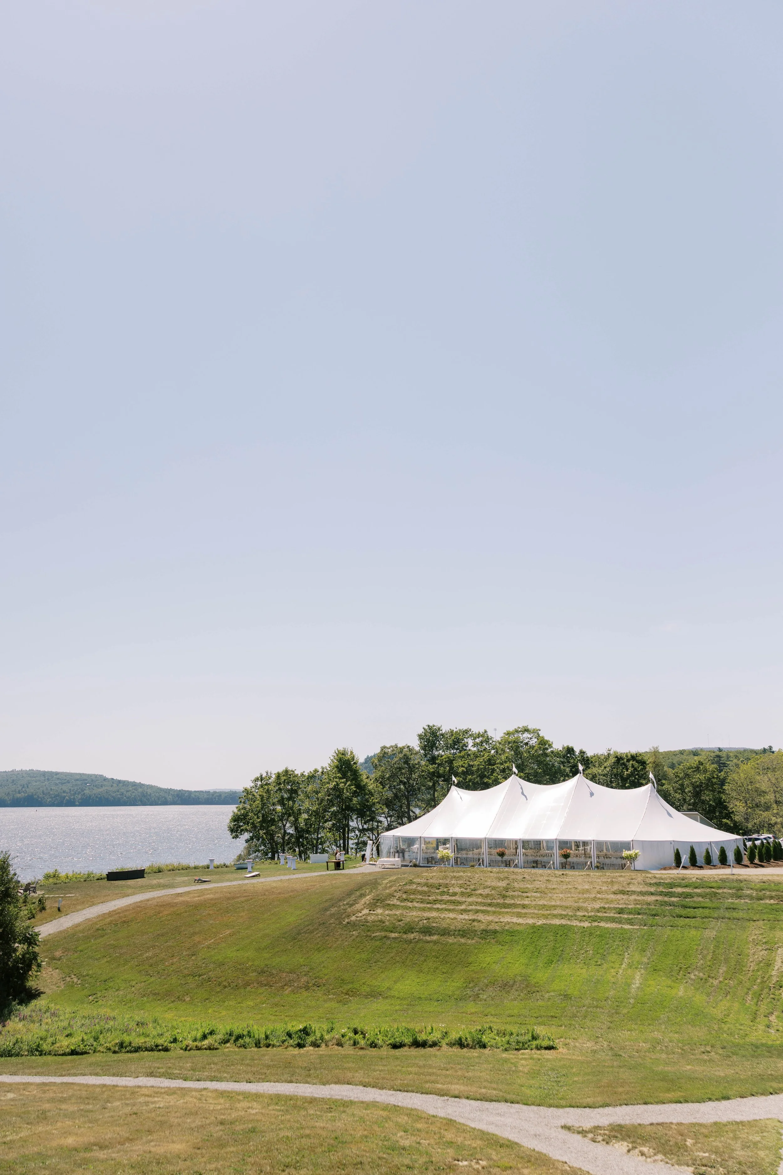 Wedding Venue in Maine by the water. A large white event tent set up on a grassy hill near a lake, with trees in the background and a clear sky overhead.