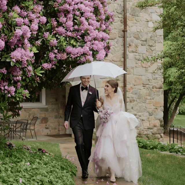 Wedding videographer in MA. Bride and groom walking outside in the rain under a white umbrella, beside a flowering pink tree, with a stone building in the background. wedding venue in MA . 