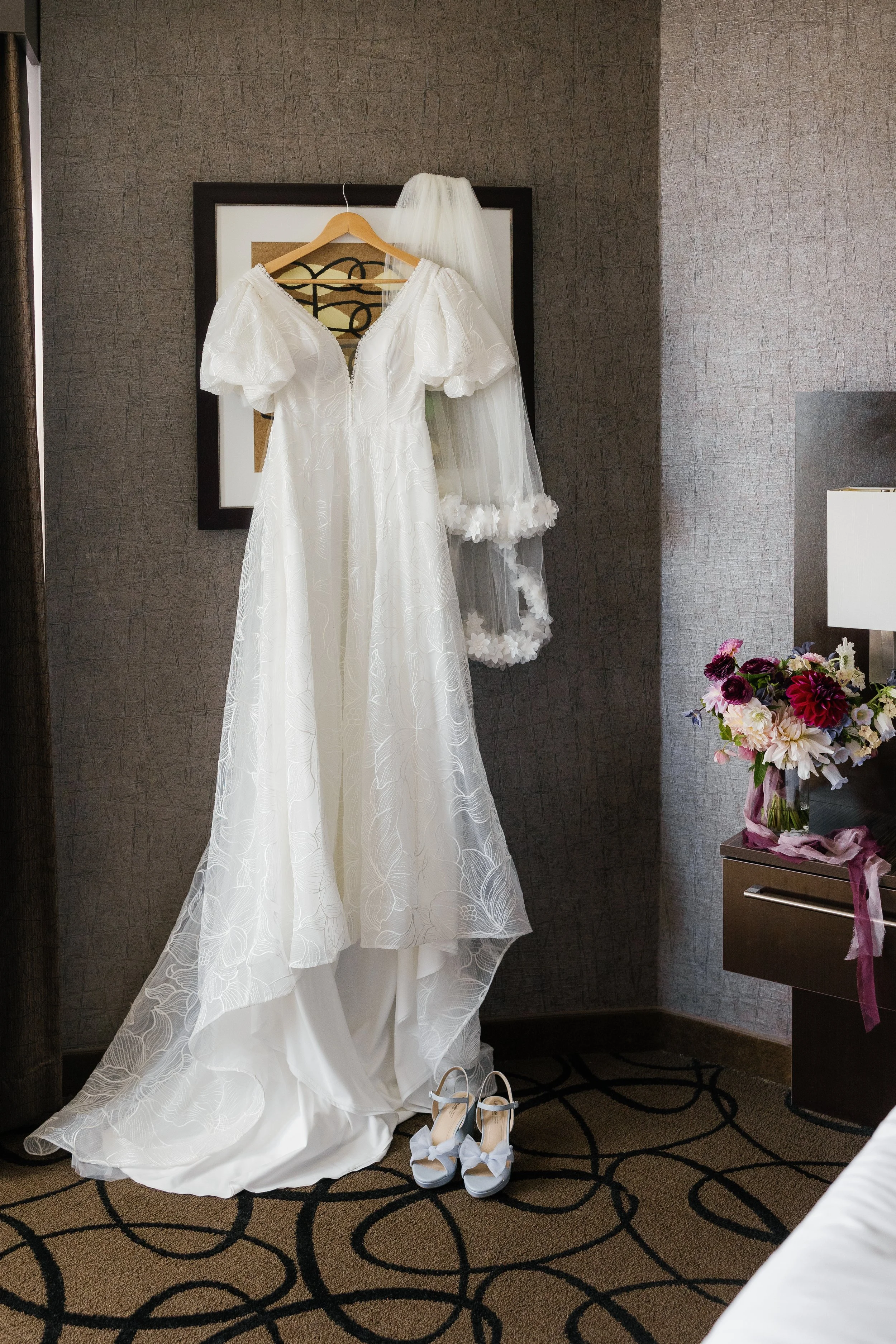 A white wedding dress hangs on a wooden hanger, with white bridal shoes placed below it, and a white veil draped behind the dress in a hotel room.