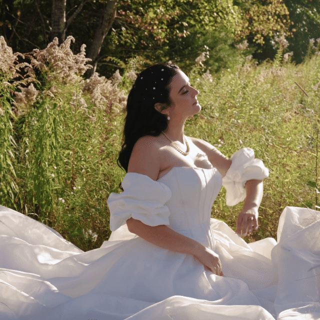 Wedding dress, wedding fun, wedding traditions all captured by the wedding videographer. A bride in a white strapless ball gown standing outdoors among tall grass and trees, with sunlight illuminating her face and hair.
