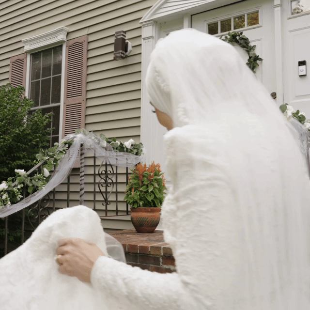 Palestinian Wedding, A woman dressed in white gently petting a fluffy white dog on a front porch decorated with flower pots and greenery. Wedding Venue at LakeView in Massachusetts. Wedding Videographers were Lasting Legacy Films