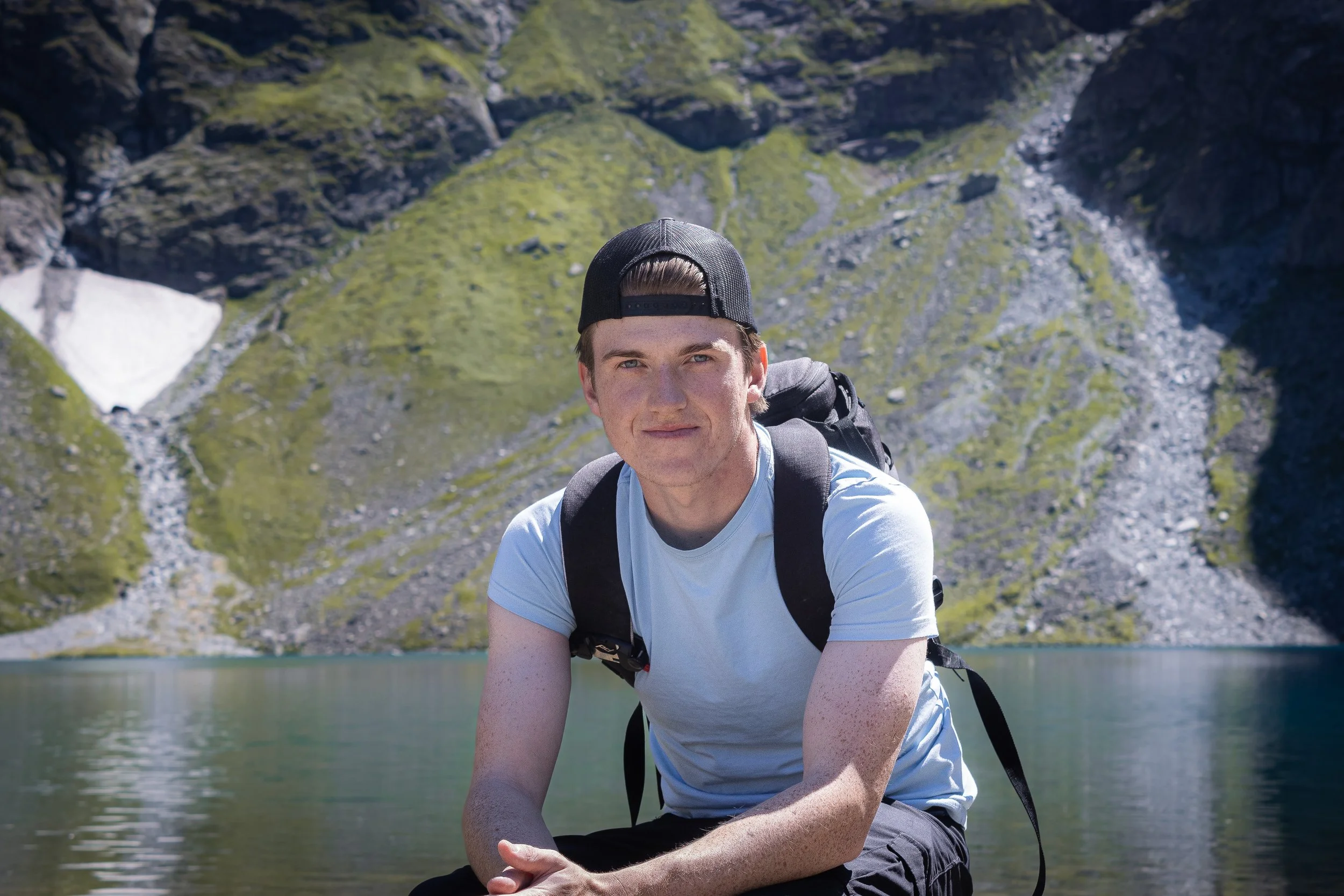A young man in a white T-shirt and black cap taking a break during a hike near a mountain lake with lush green slopes and rocky cliffs in the background.