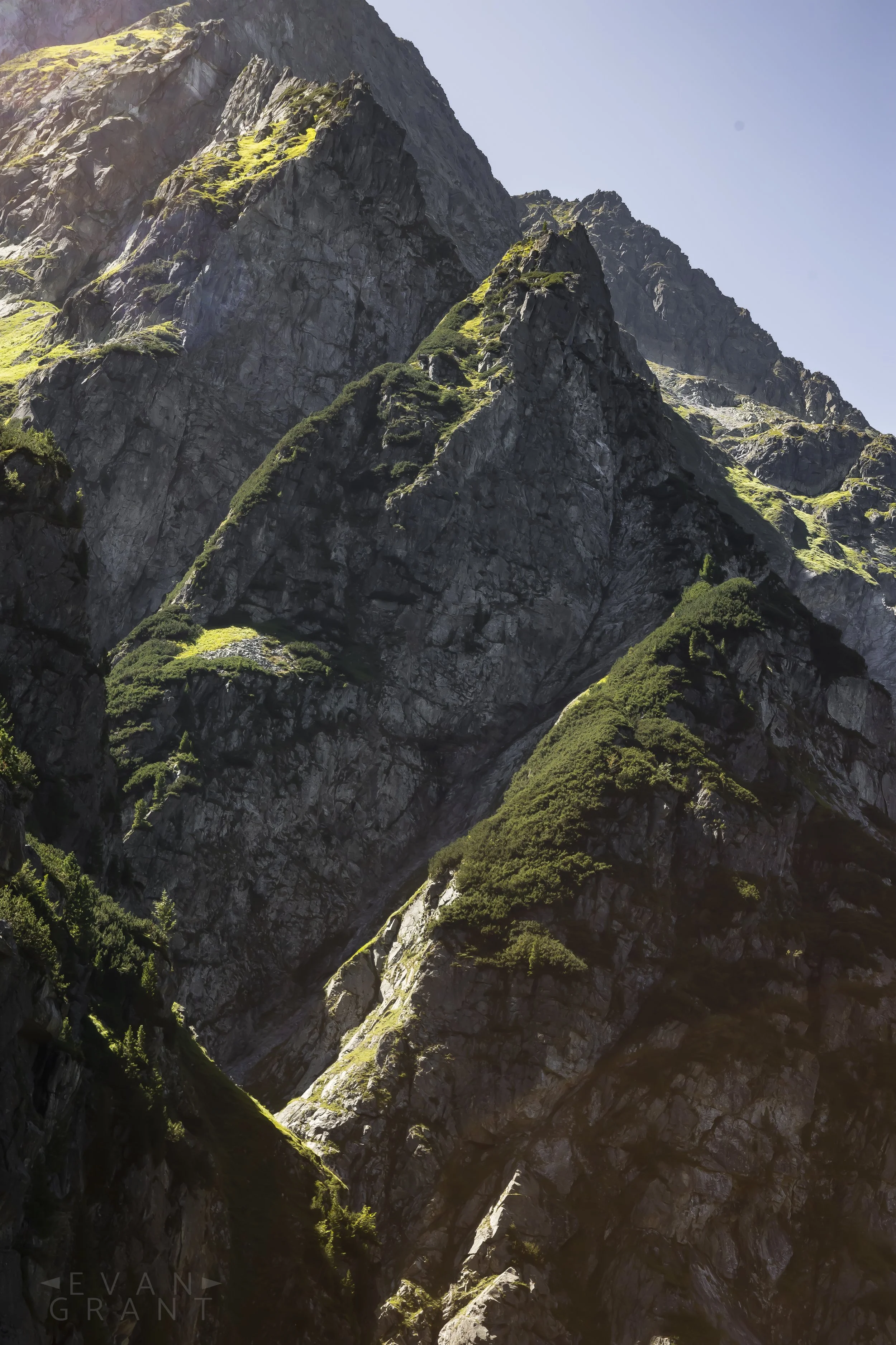 Tall rugged mountain peaks with patches of green vegetation under a clear blue sky.