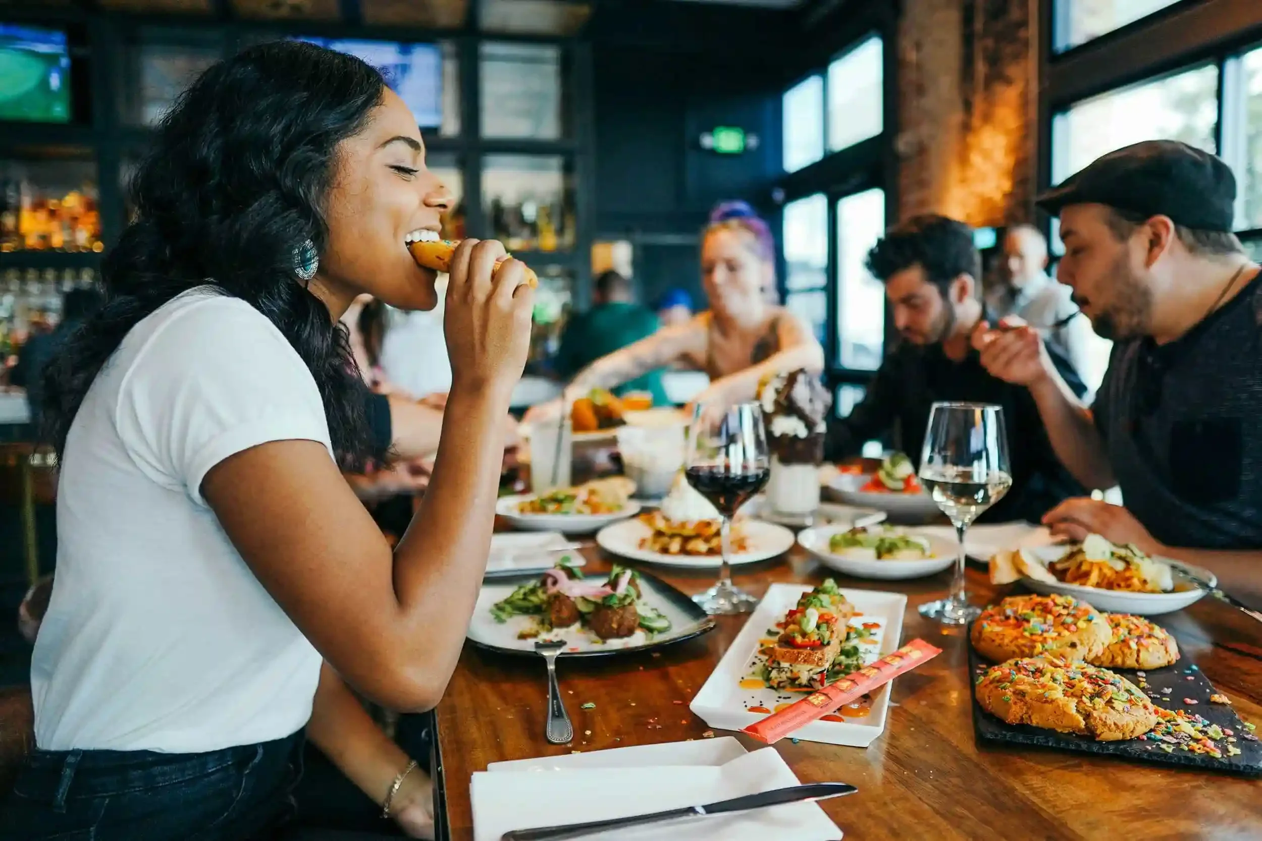 People enjoy food and drinks together at a lively restaurant, sharing plates and conversation around a wooden table.
