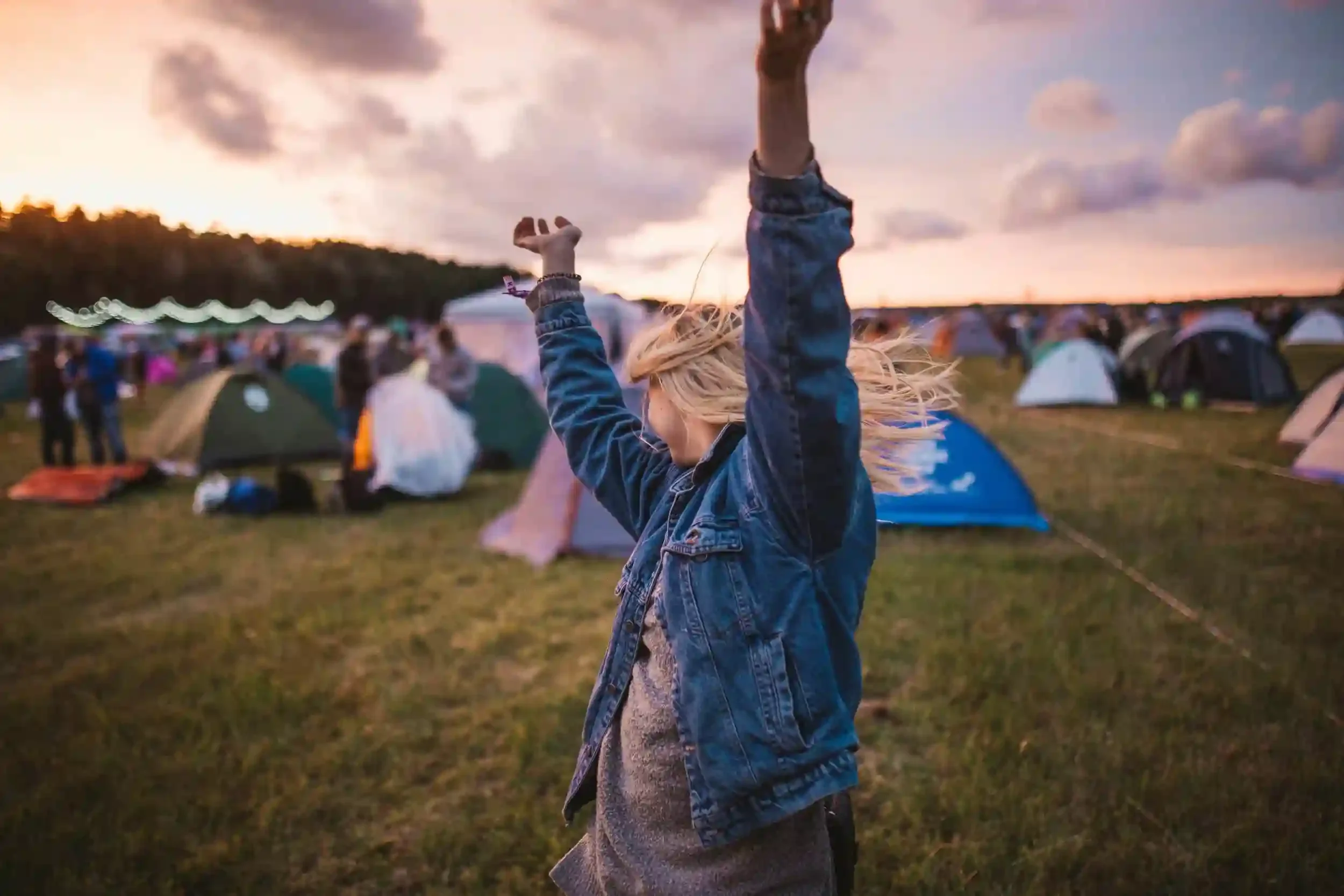 A woman with arms raised enjoys a lively outdoor camping event at sunset, surrounded by tents and people.