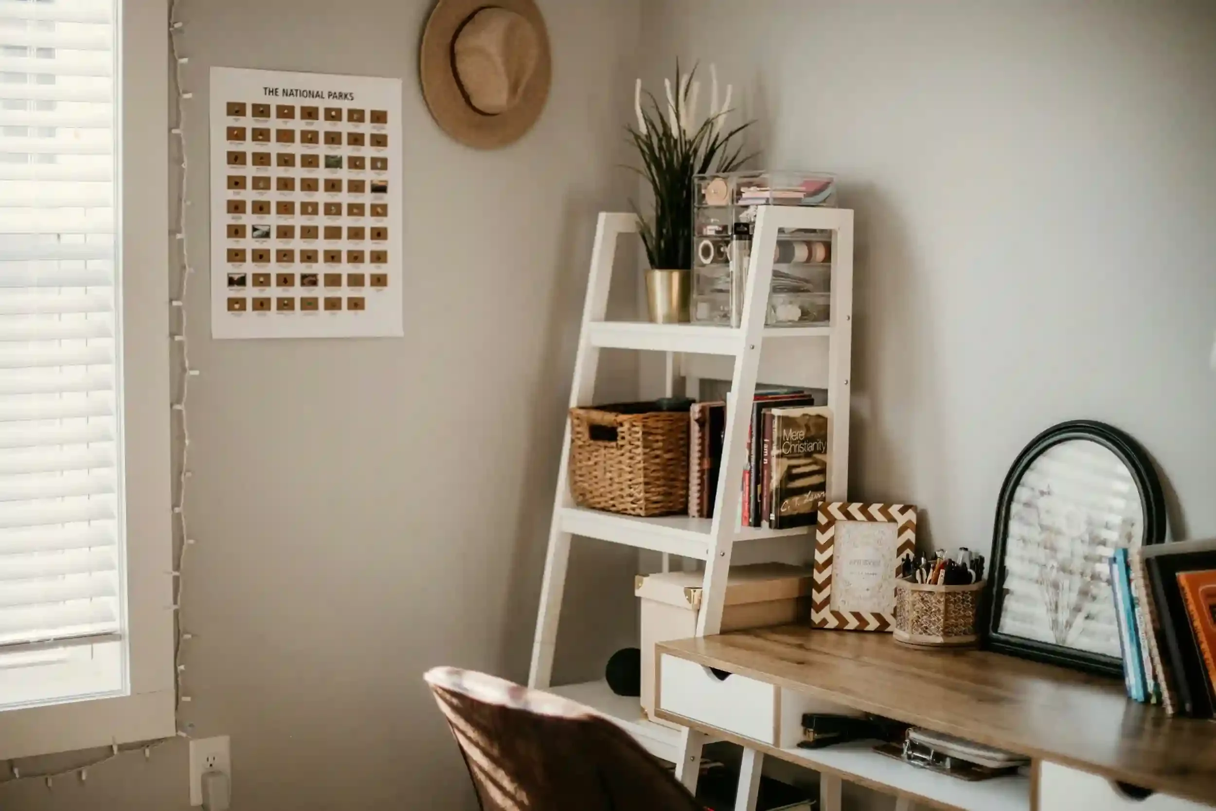 A cozy home office with a desk, shelves, basket storage, plants, a wall poster, and natural light from a window.