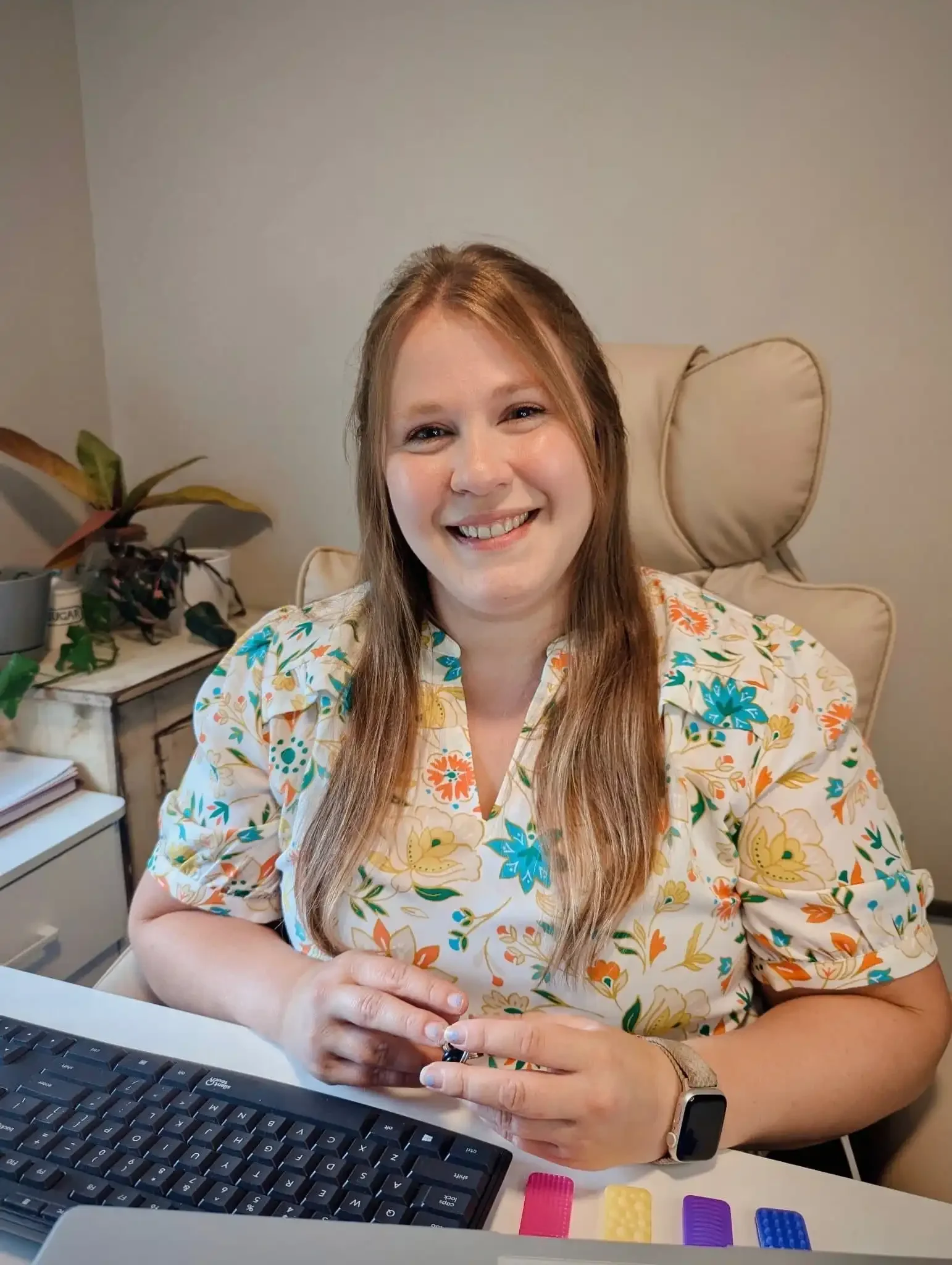 Bethany Valenti in a floral blouse smiles at a desk with a keyboard, plants, and office supplies visible nearby.