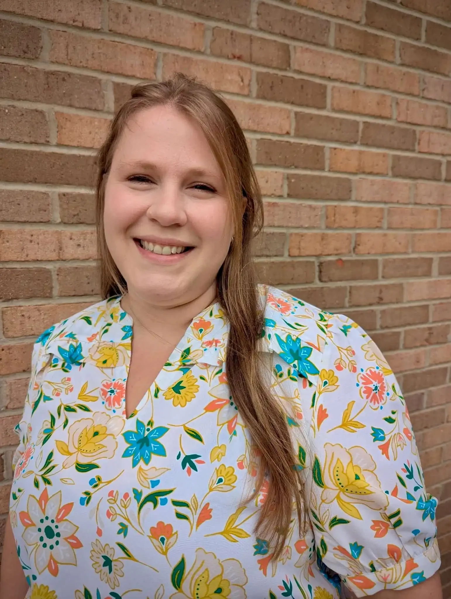 Smiling Bethany Valenti wears a colorful floral blouse and stands in front of a brick wall.