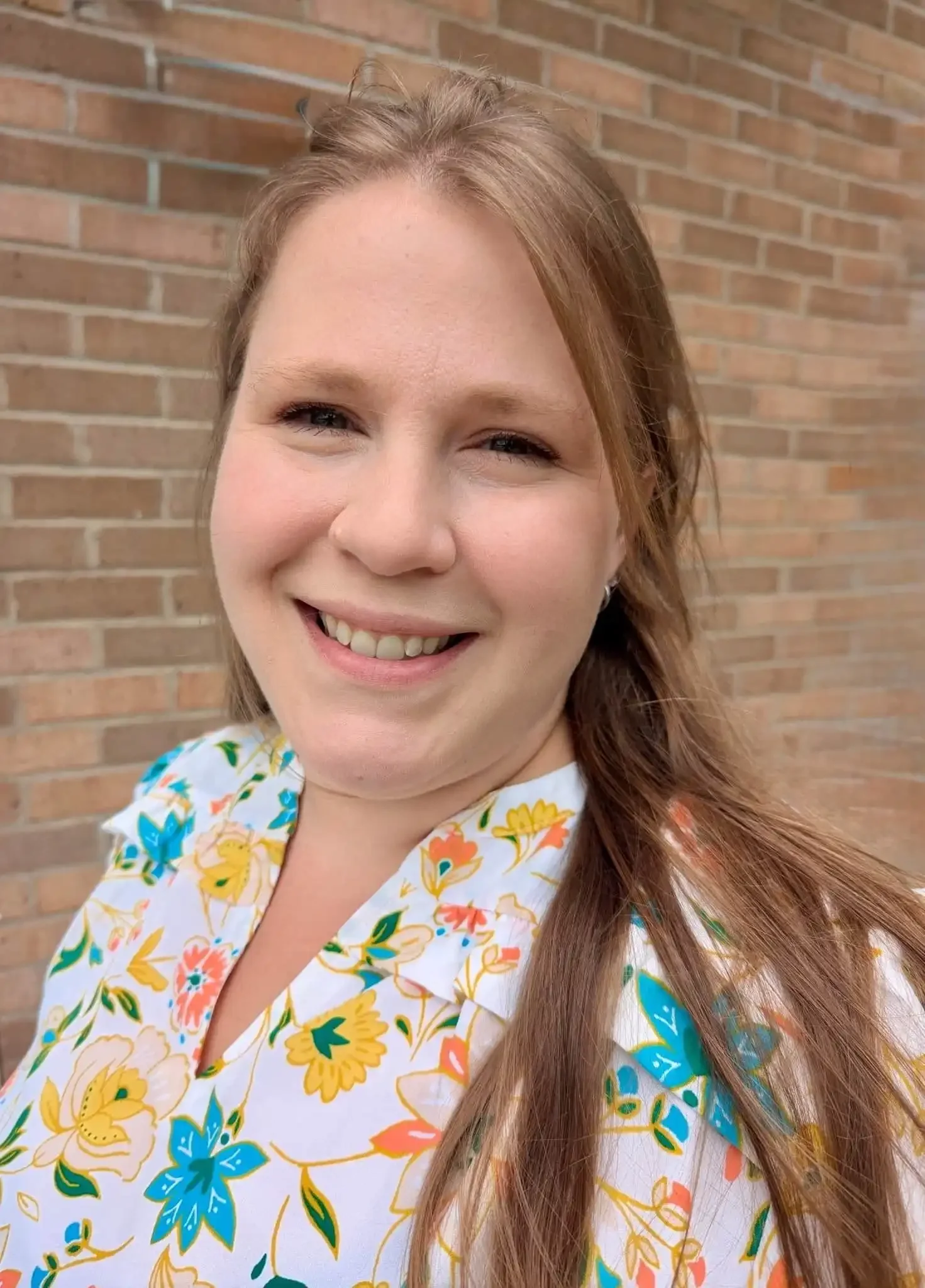 Bethany Valenti smiles, wearing a floral top, standing in front of a brick wall.