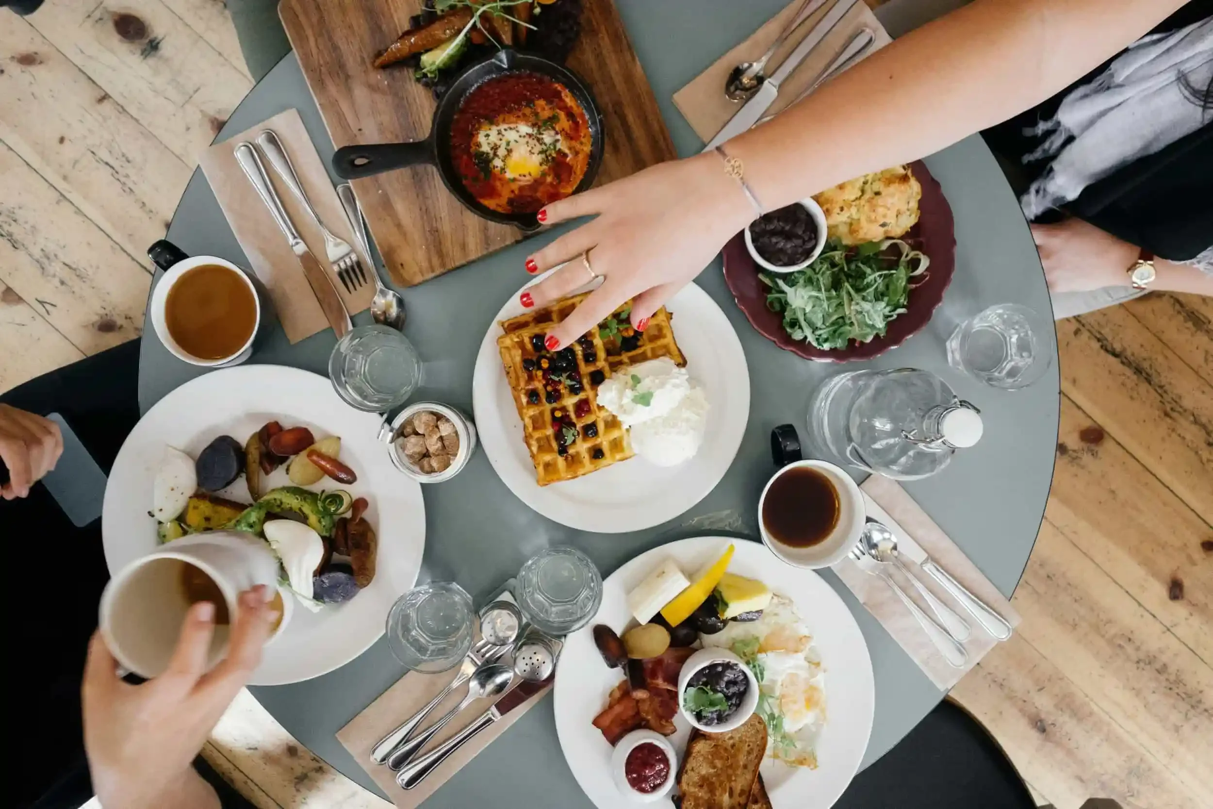 Overhead view of a breakfast table with waffles, eggs, coffee, and hands reaching for food on a round green table.
