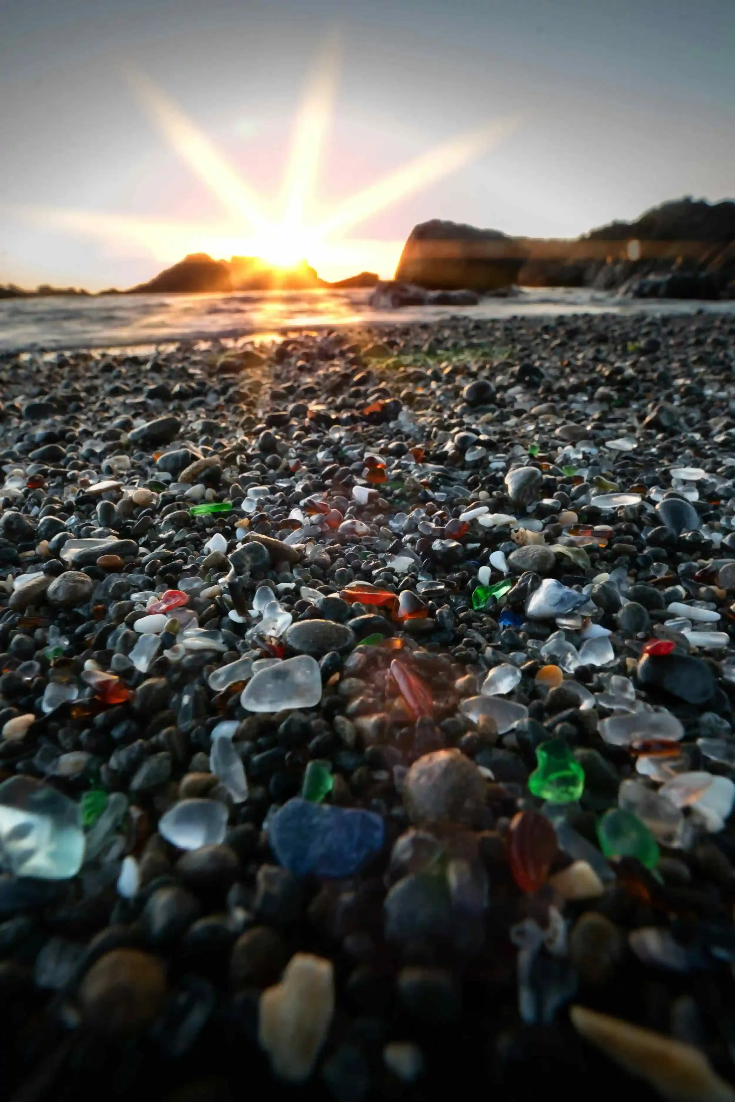 Colorful sea glass and pebbles on a beach at sunset, with the sun shining brightly over distant rocks and the ocean in the background.