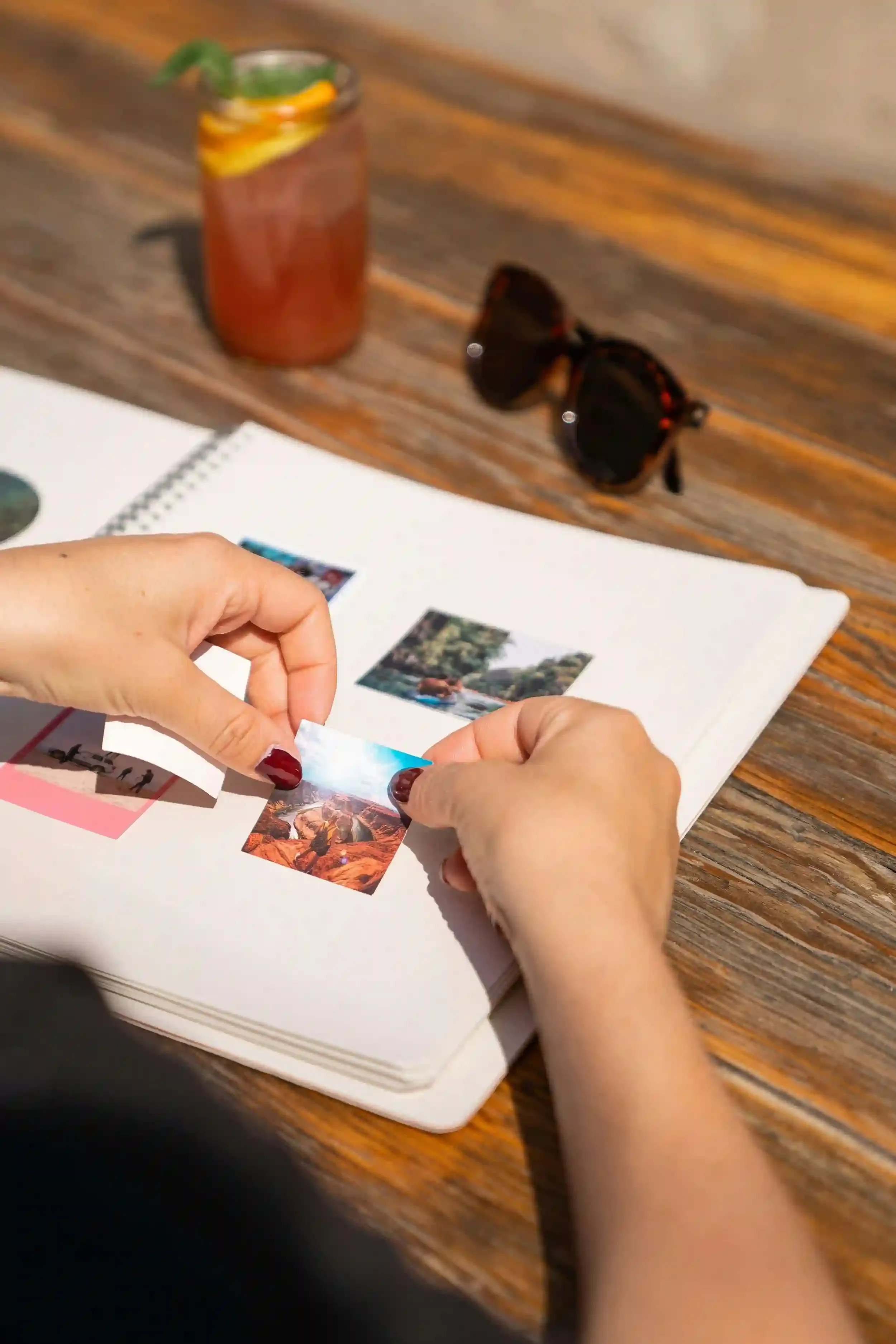 A person arranges small photos in a scrapbook on a wooden table with sunglasses and a drink nearby.