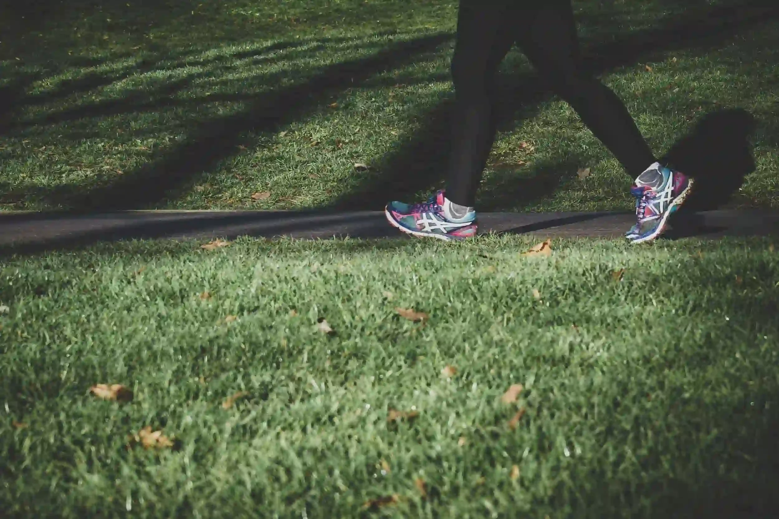 A person wearing black leggings and colorful running shoes walks on a path bordered by green grass.