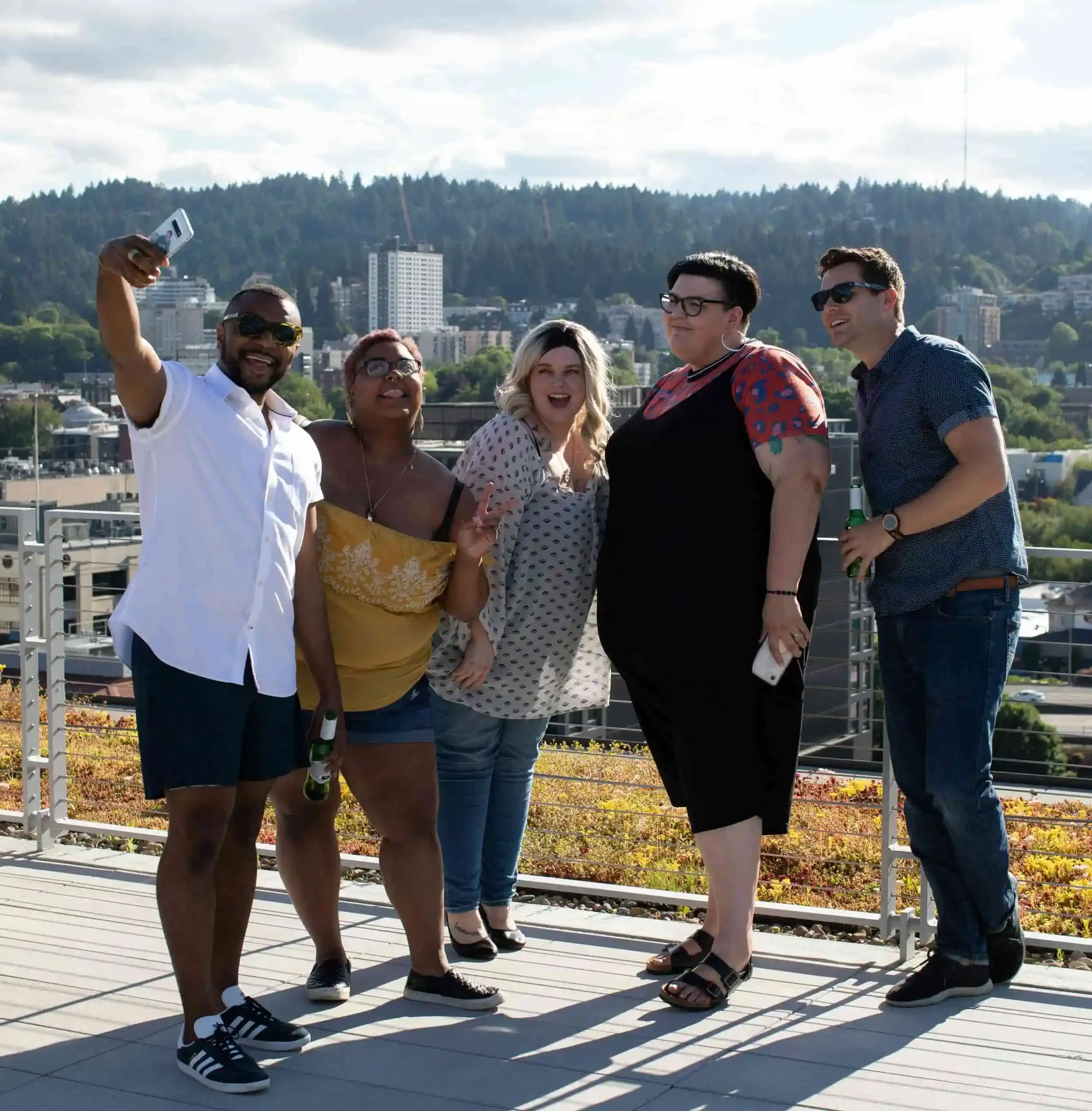 Five people smiling and taking a group selfie on a rooftop with a city and forested hills in the background.