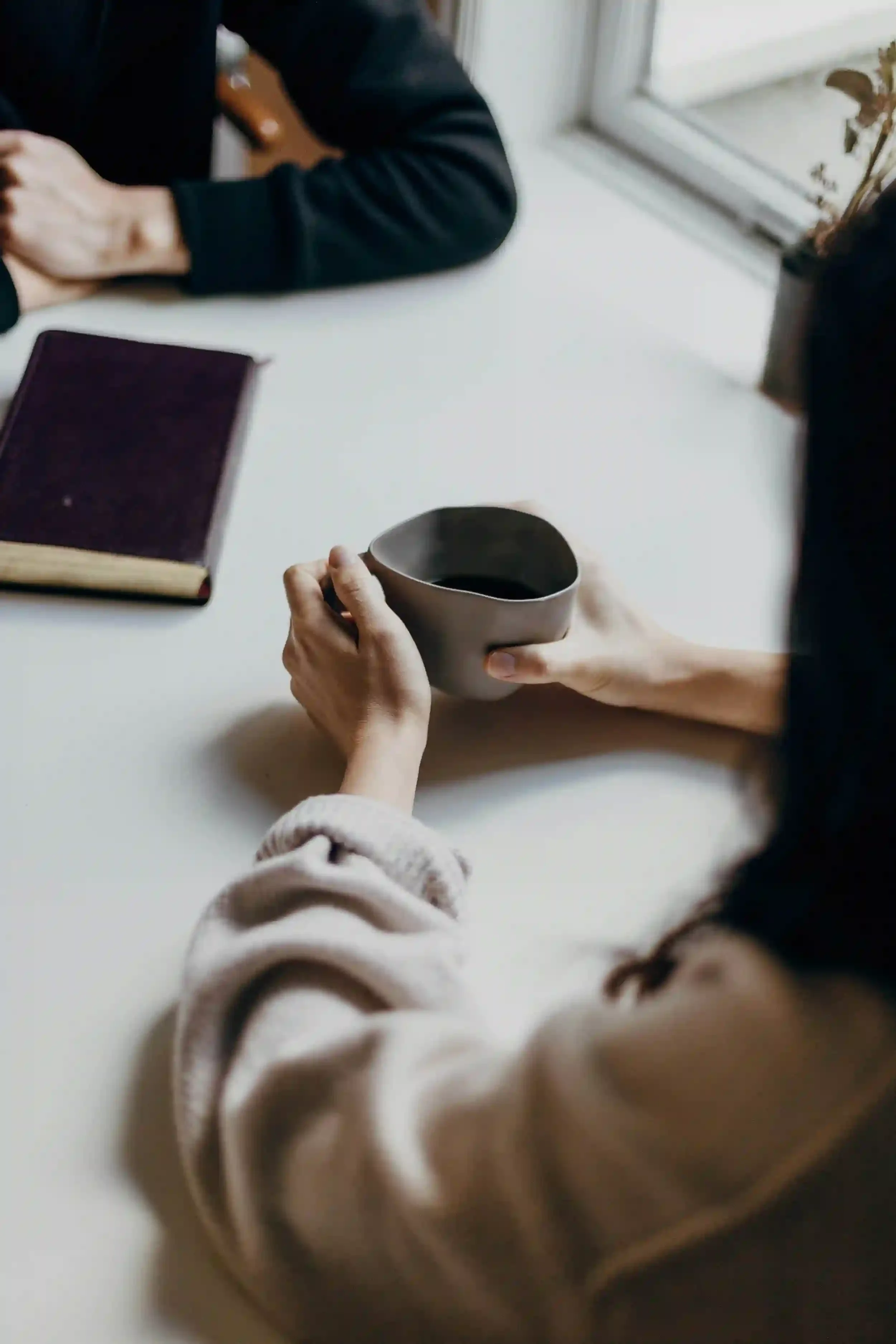A person holding a mug sits at a white table with a closed book and another person’s hands visible nearby.