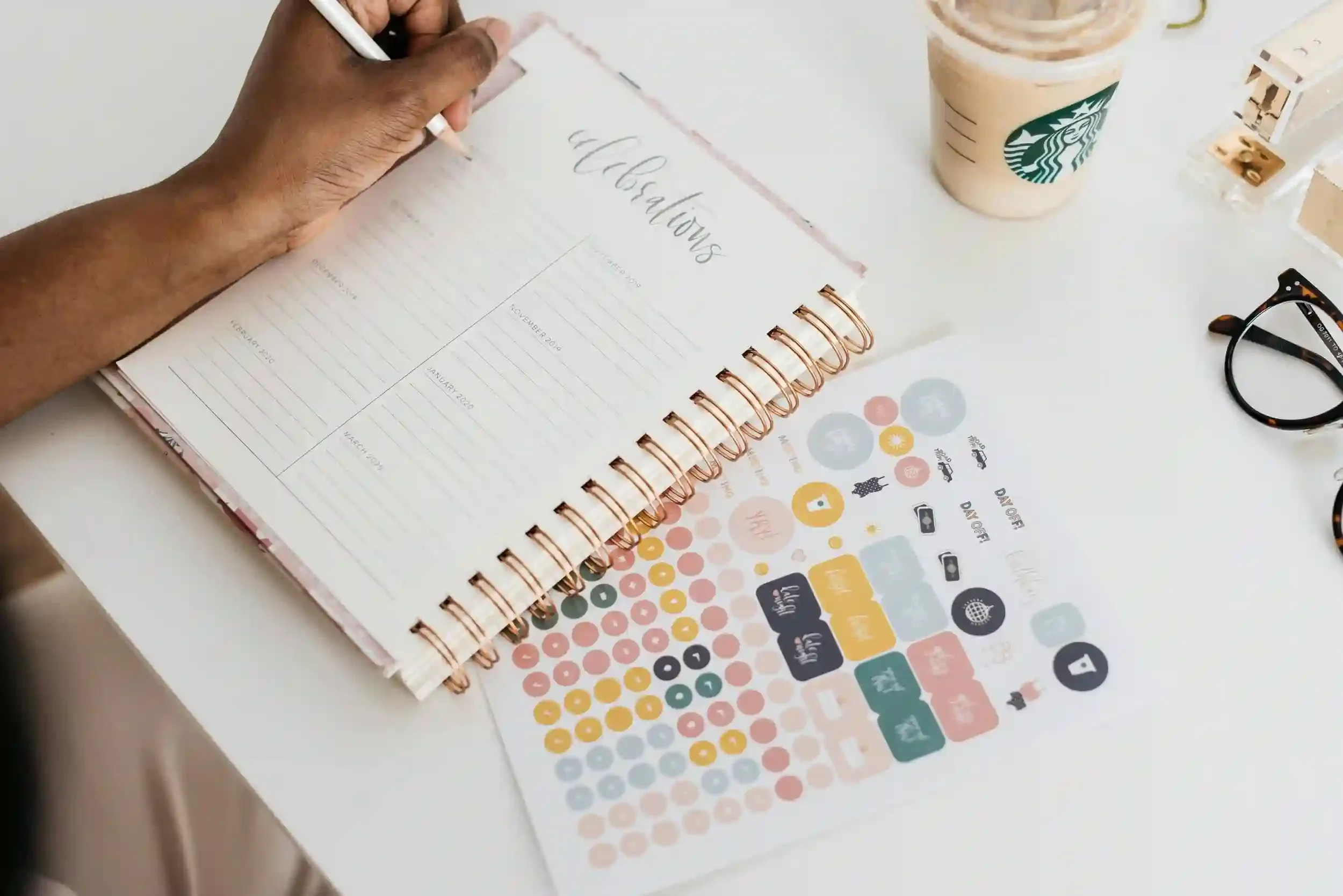 A person writes in a planner labeled celebrations with colorful stickers and an iced Starbucks drink on the desk.