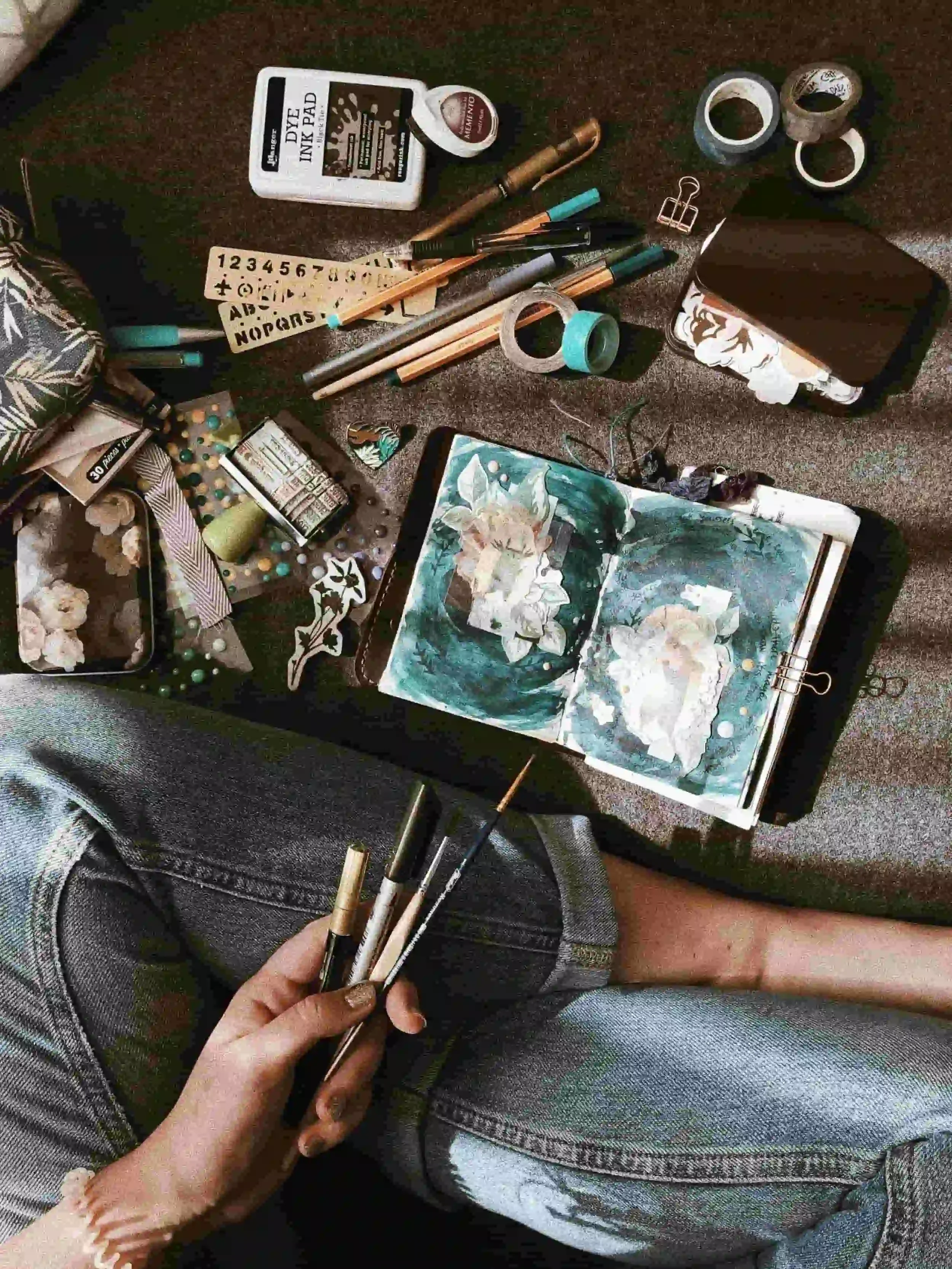 Person sitting cross-legged with art supplies, brushes, and an open sketchbook featuring a watercolor painting on their lap.