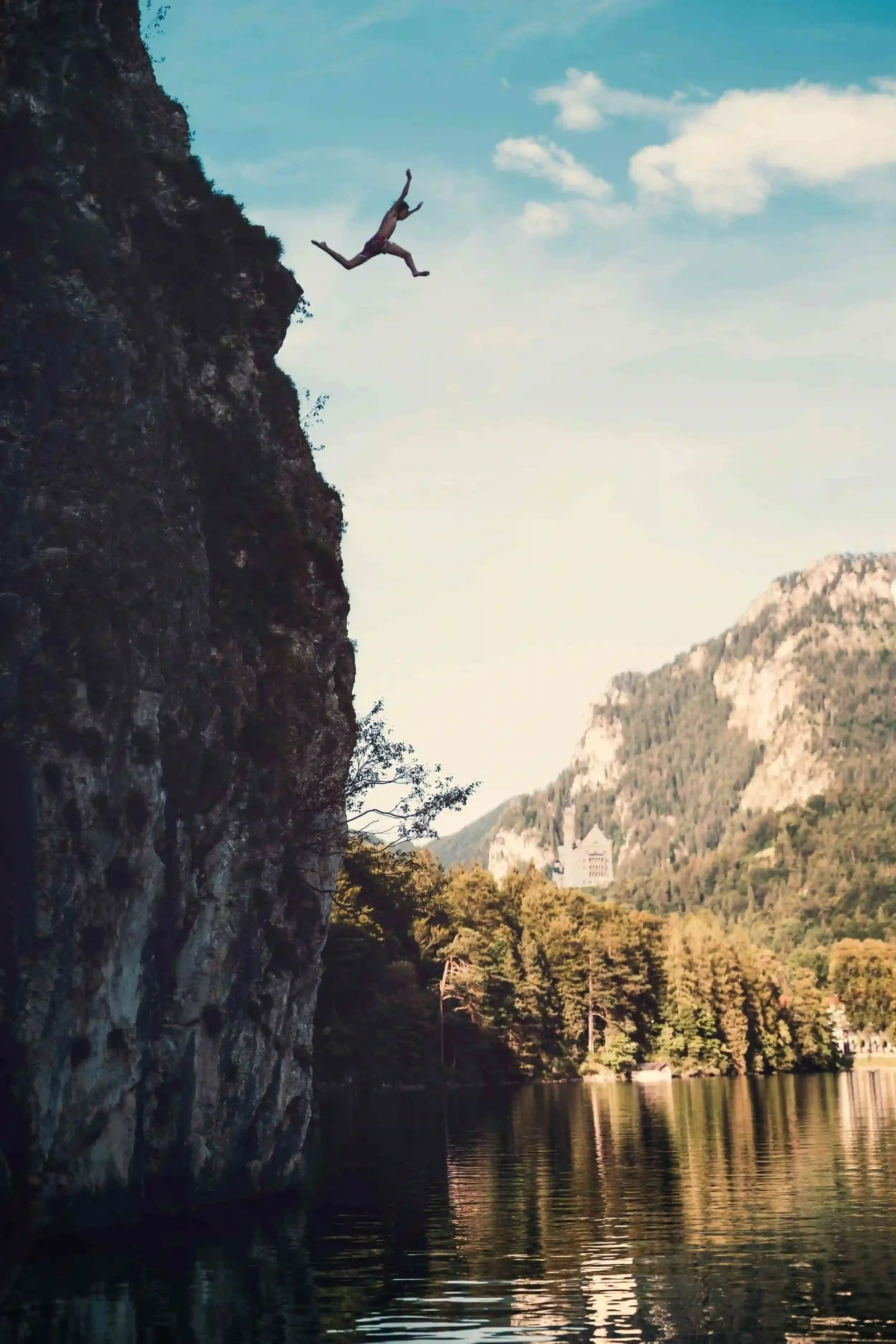 A person jumps off a steep cliff into a calm lake, surrounded by trees and mountains under a blue sky.