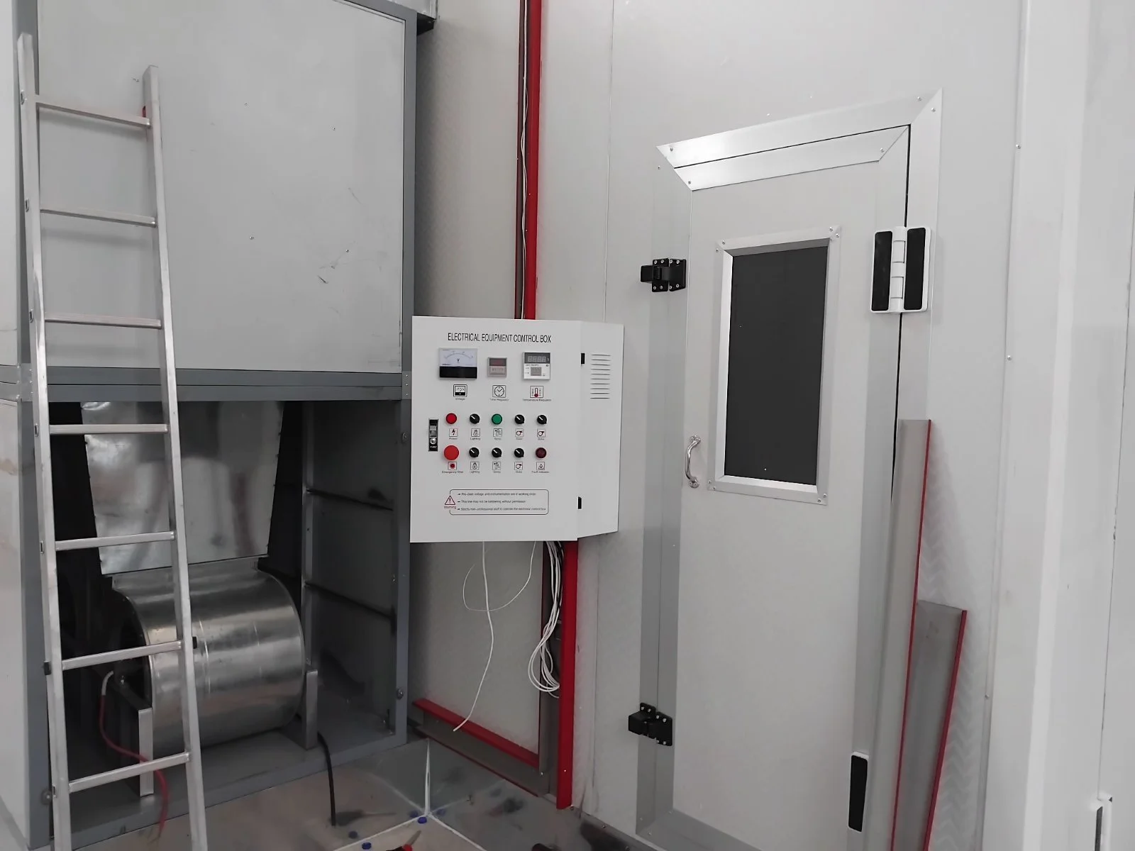 Ein Raum mit einer Metallleiter, einem Schaltschrank, einem roten Kabel, einem schwarzen Gehäuse und einer Tür mit Sichtfenster