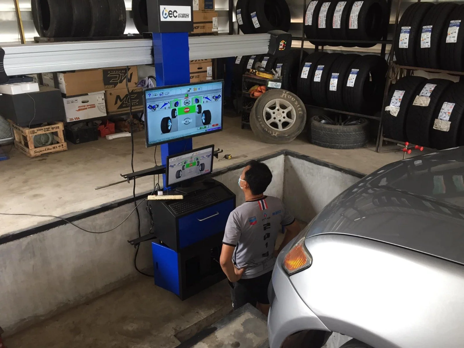 Ein Mechaniker in einer Werkstatt arbeitet an einem Computerbildschirm, der eine Fahrzeugsteuerung oder -überwachung zeigt, während er vor einem geöffneten Grube steht, mit Reifen und Autoteilen im Hintergrund.