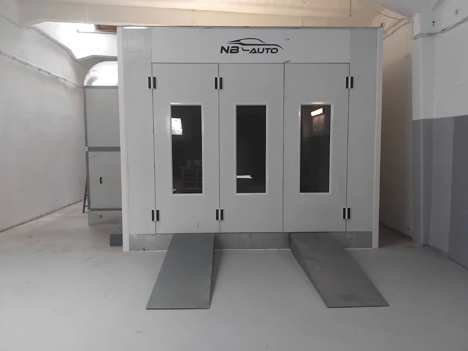 Eine Grau metallenisierte Fahrzeugwaschbox mit drei Türen mit Glaspanelen, auf der sich die Aufschrift 'NB-AUTO' befindet, steht in einem Parkhaus.