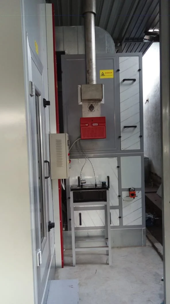 Industrielle Abzugshaube mit Dunstabzug, Kontrollpanelen, eine kleine Arbeitsstation mit Wasserhahn, und Sicherheitsschilder in einer Werkstatt oder Fabrik.