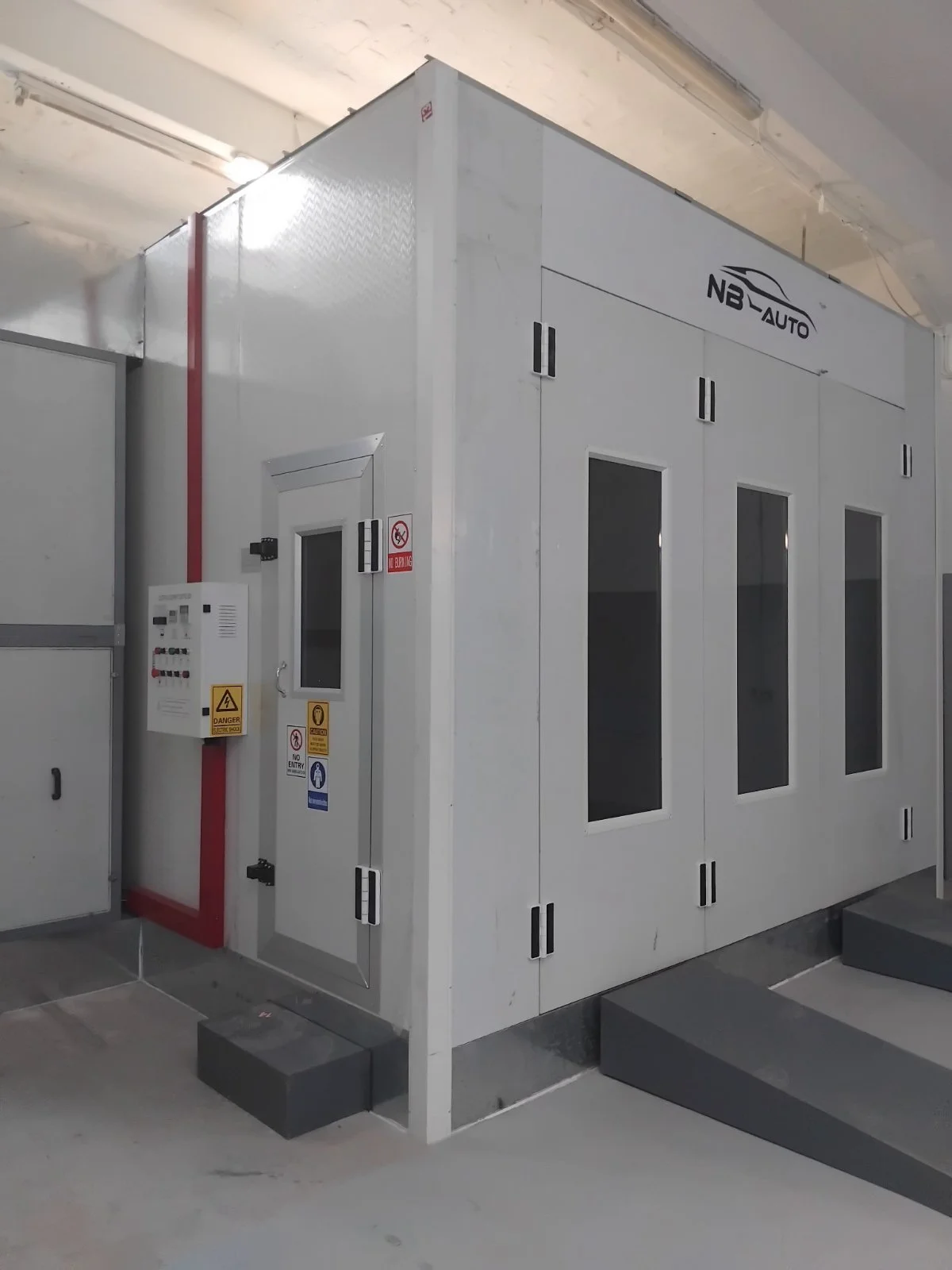 In einem Keller befindet sich eine industrielle Klimaanlage oder Kälteanlage in einem weißen Gehäuse mit vier quadratischen Lüftungsschlitzen, einem kleinen Türchen und Warnschildern, darunter ein Handfeuerlöscher- und Gefahrenschild.