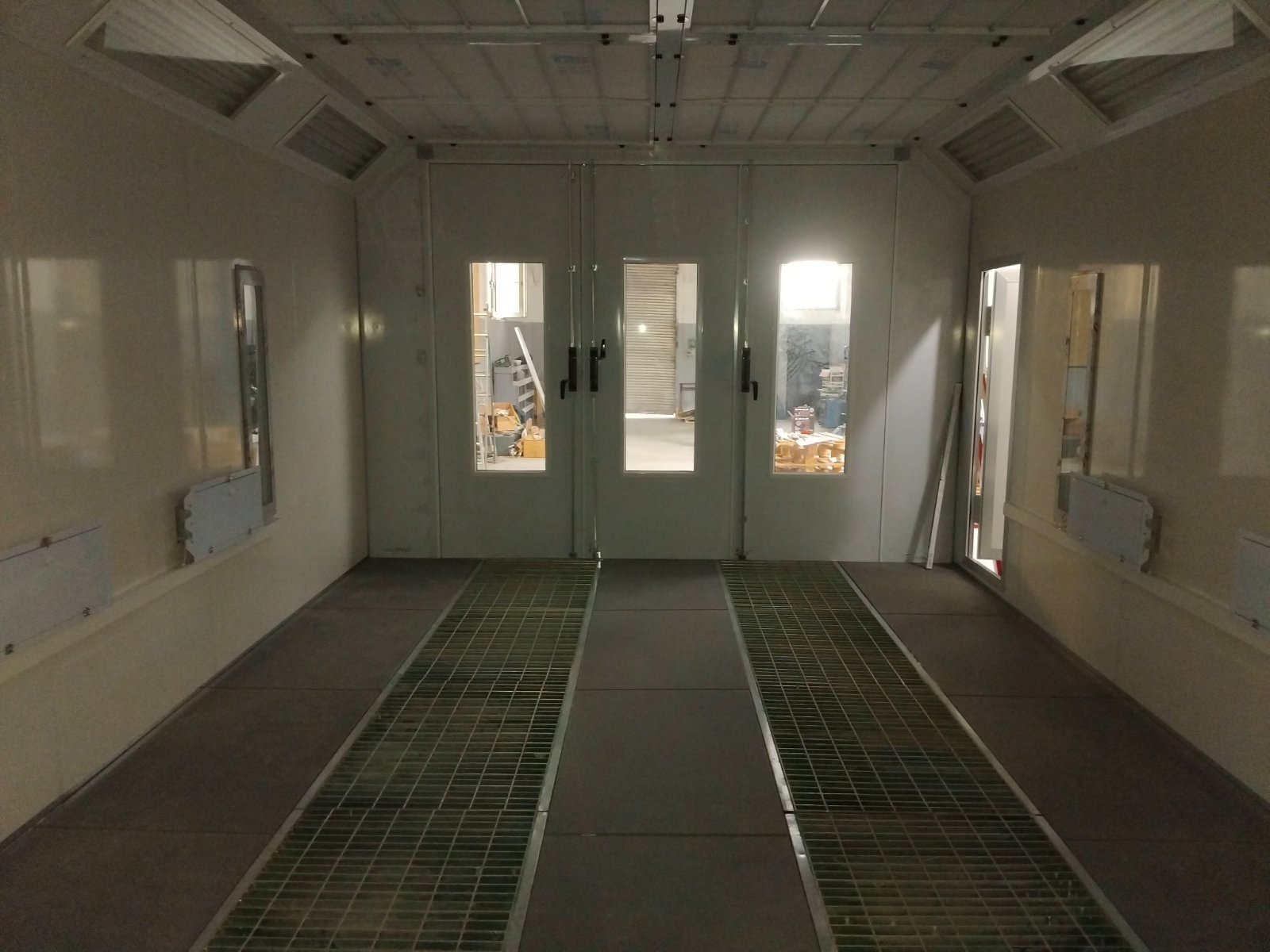 Ein Raum mit grauen Türen mit Fenstern, die in eine Werkstatt oder Lagerbereich führen, mit Gitterboden und einigen Baumaterialien.