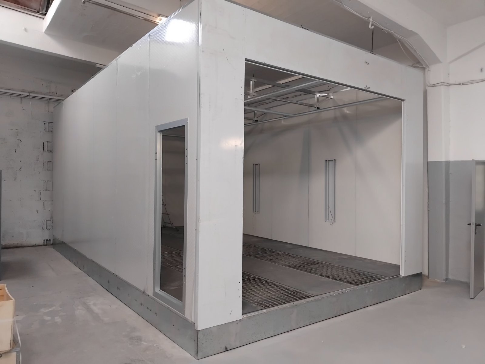 Ein leerer, rechteckiger Raum mit weißverkleideten Wänden, einem offenen Eingang und einigen Metallgittern auf dem Boden, wahrscheinlich im Bau oder in der Renovierung.