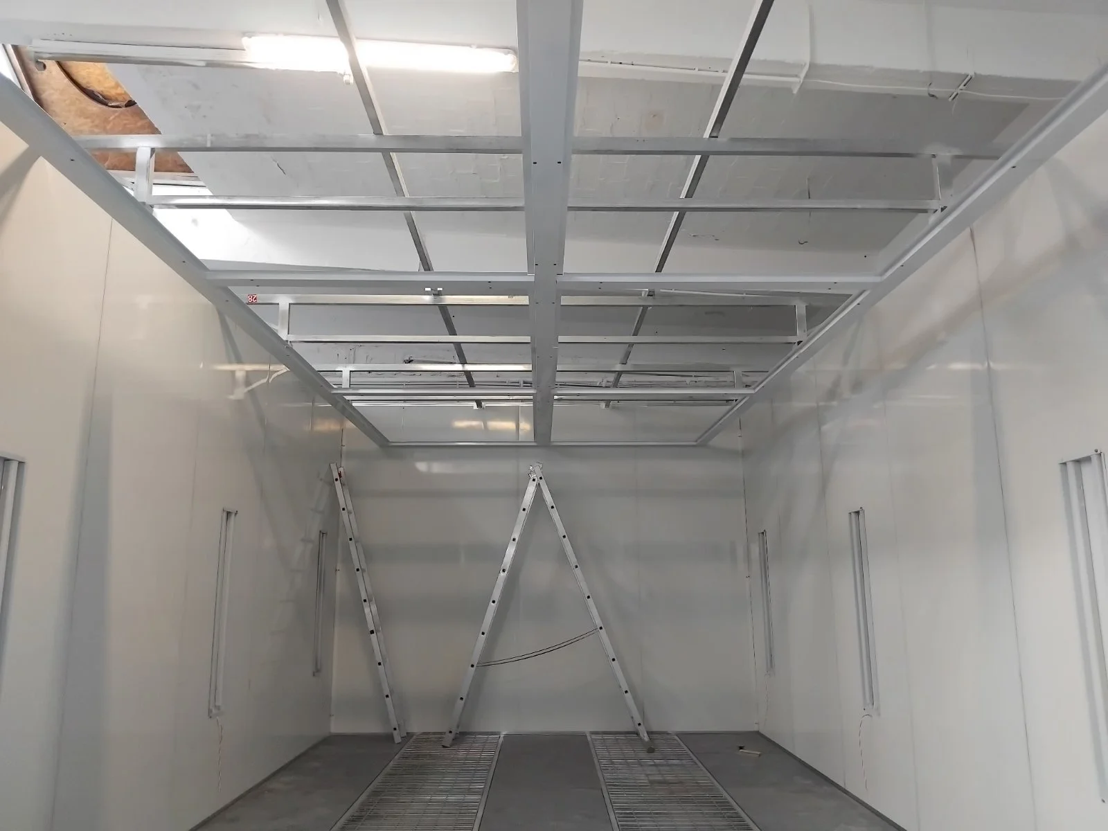 Innenraum eines Raums unter Ausbau, mit weißen Wänden, Metallrahmen an der Decke, zwei Leitern und Metallgitter auf dem Boden.