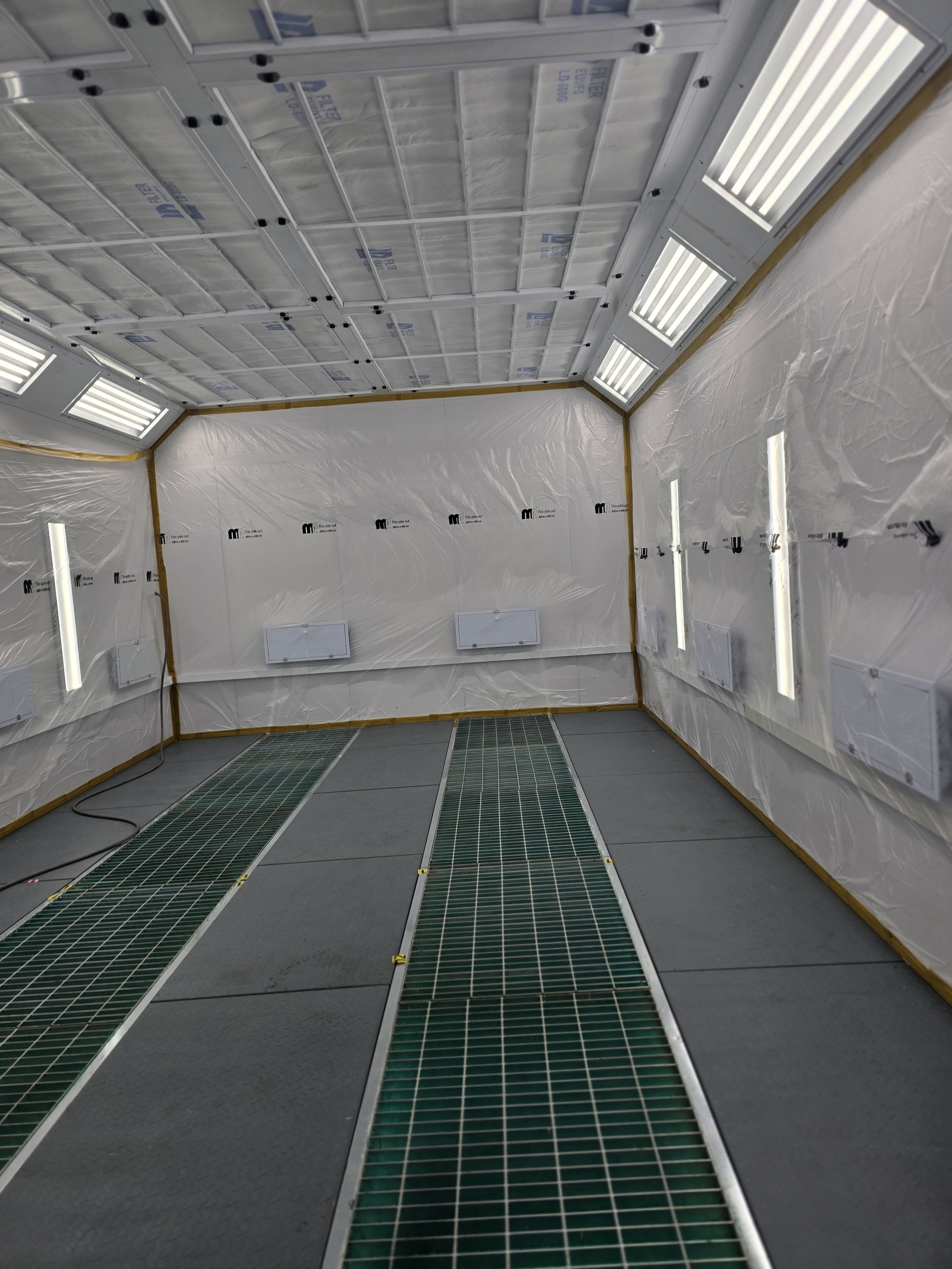 Innenraum eines Raumes mit Metallgittern auf dem Boden, weißen Wandverkleidungen mit Heizungen und Decke mit Lichtleisten, wahrscheinlich eine Lackierkabine oder ein industrieller Arbeitsraum.