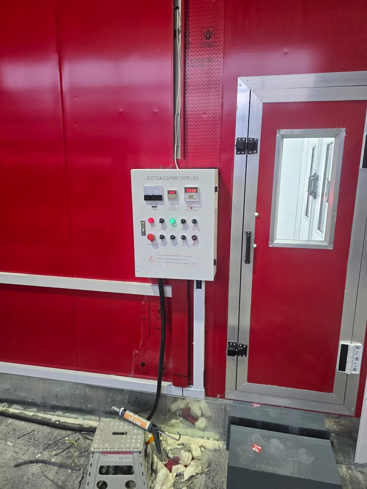 Ein rotes Raum mit einer schwarzen Steuerbox an der Wand, die verschiedene Knöpfe und Anzeigen hat, sowie einem kleinen Fenster auf dem Türrahmen.