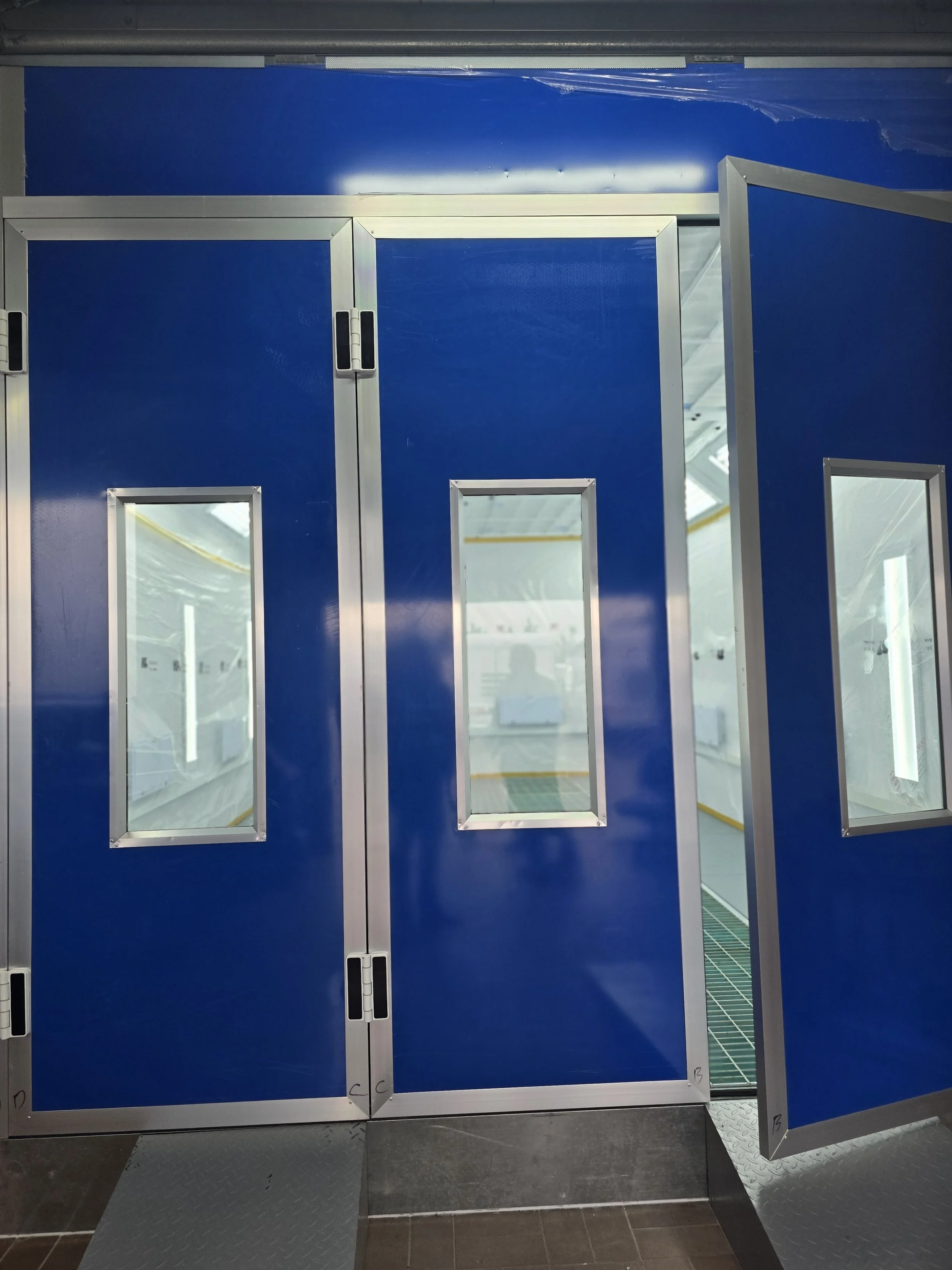 Blaue Industrietür mit kleinen Fenstern, eine Tür ist offen, im Inneren ist eine ausgeleuchtete Fertigungsstätte sichtbar.